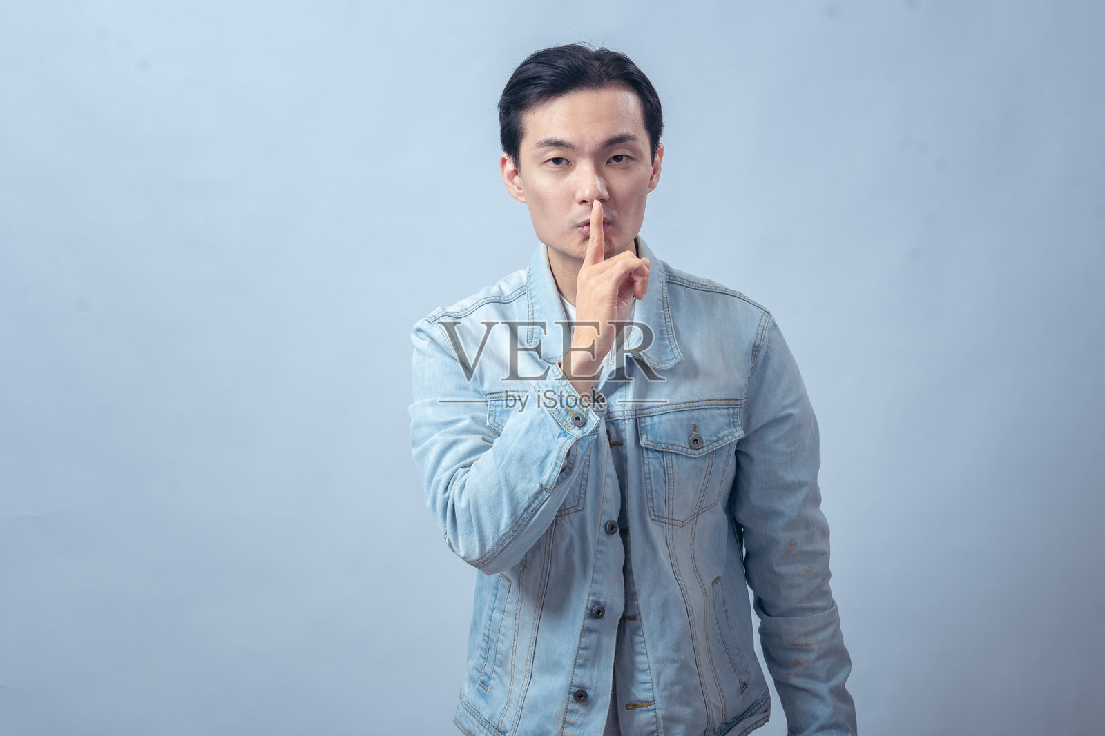 一名亚洲男子身穿牛仔夹克，食指放在嘴唇上做出噤声手势，表情严肃，在影棚背景下，象征着安静、秘密或保密的概念照片摄影图片