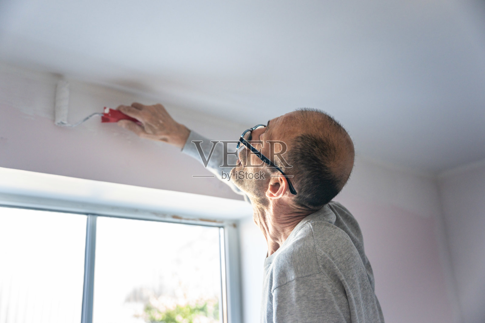 男人在自然光照下，在家里的室内用滚筒刷给靠近天花板的墙壁刷漆照片摄影图片