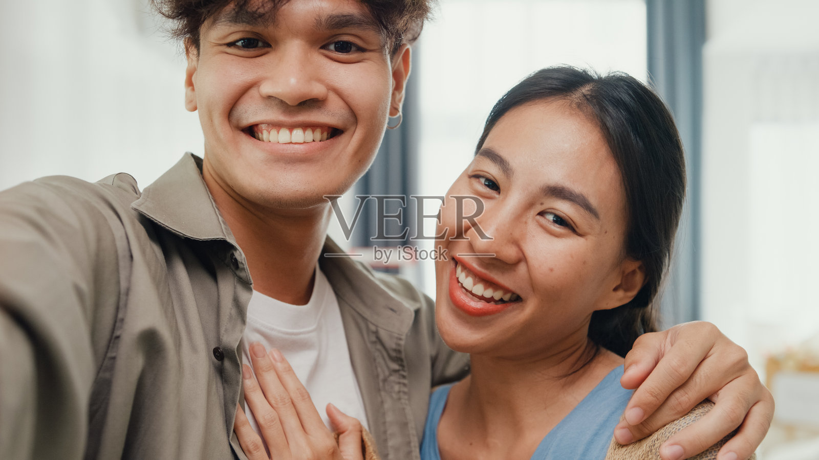 一对年轻快乐的亚裔情侣正在进行视频通话，他们看着镜头，自豪地微笑，并向客厅里的朋友们展示他们的新家。夫妻搬入新家，公寓出租与所有权。照片摄影图片