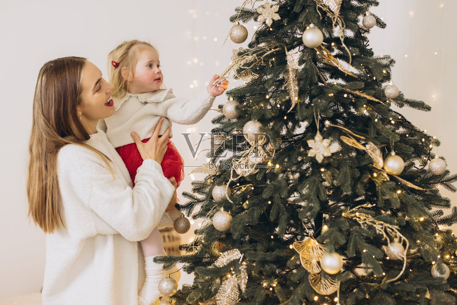 妈妈在装饰圣诞树，上面挂满了金色的装饰品，她还把女儿抱了起来。照片摄影图片