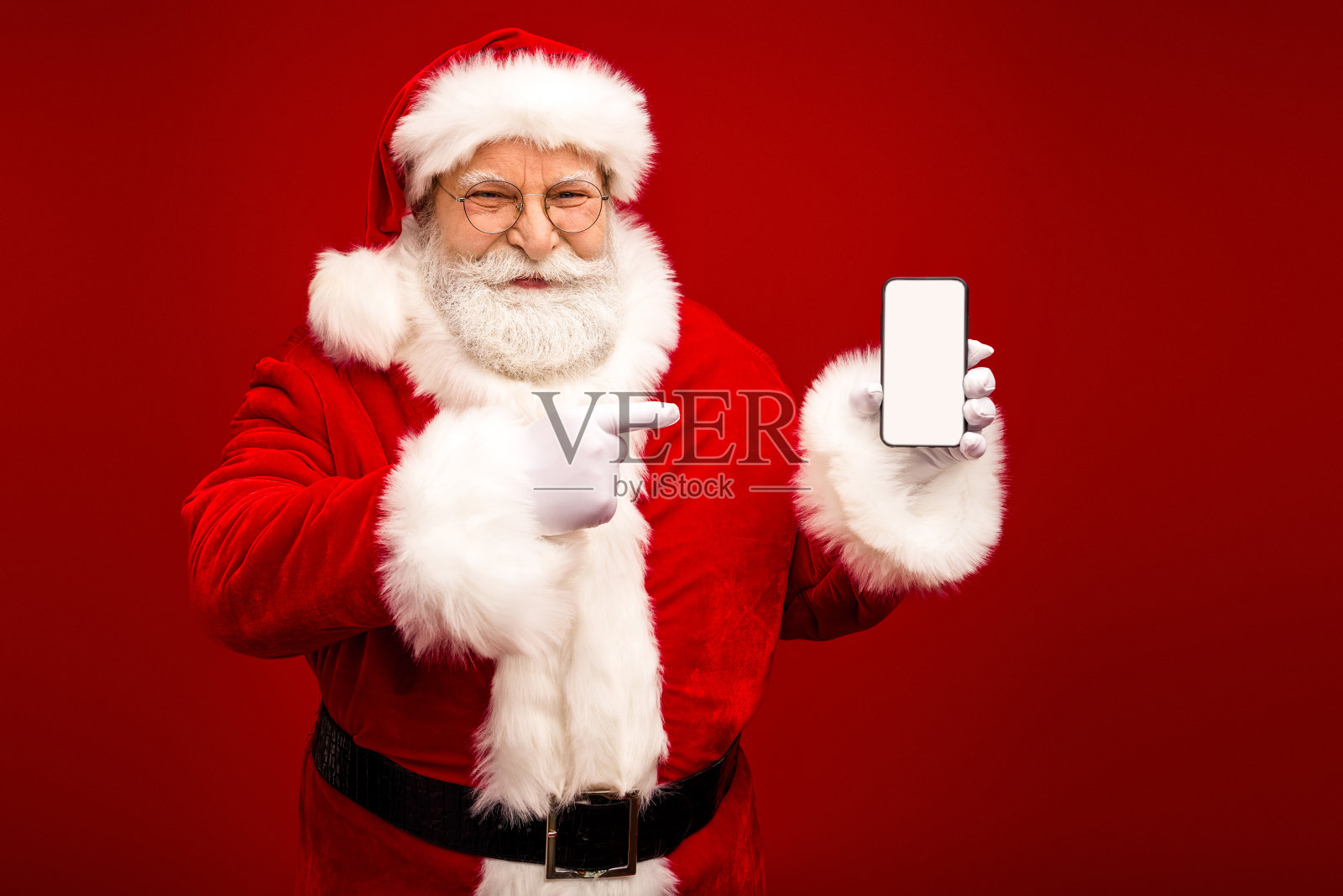 圣诞老人举着空白的智能手机屏幕，在明亮的红色背景前指向它，用于圣诞主题的素材图片照片摄影图片