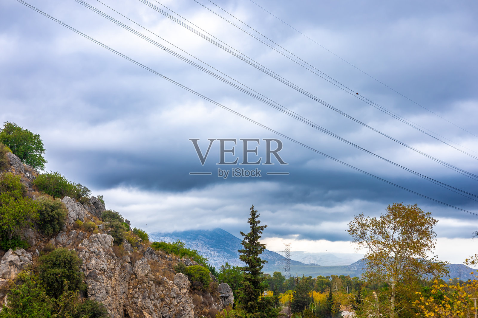 电线横跨多云的天空，下方是岩石遍布、稀疏树木点缀、远山隐现的景观，展现了与电力的连接。照片摄影图片