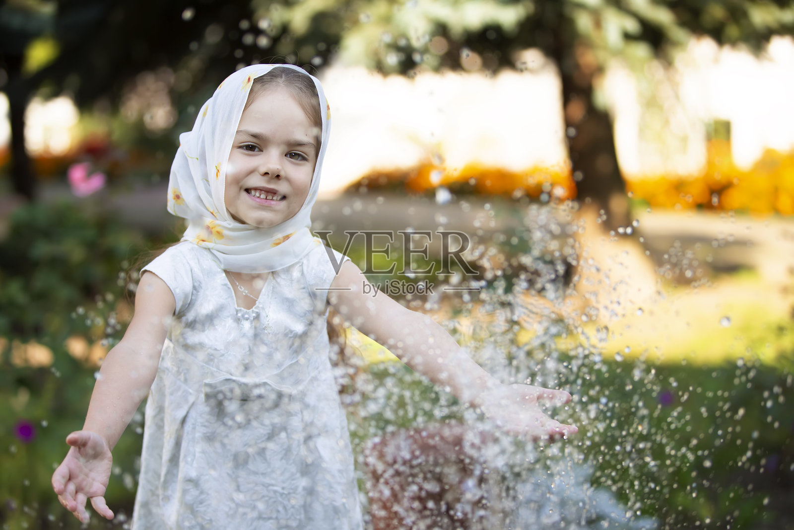 小女孩在阳光明媚的日子里，戴着一条浅色围巾，在花园里开心地泼水玩耍。照片摄影图片
