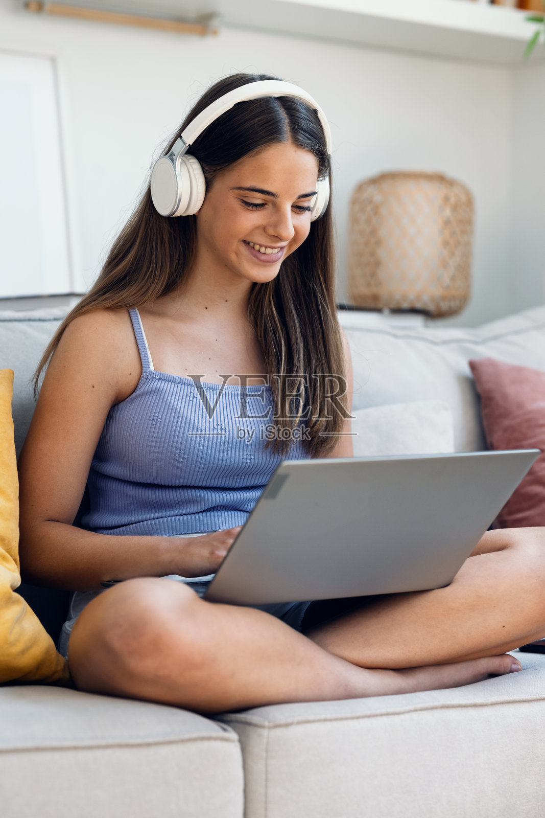 一位美丽的女孩在家中坐在沙发上，戴着耳机听着音乐，同时使用笔记本电脑工作。照片摄影图片
