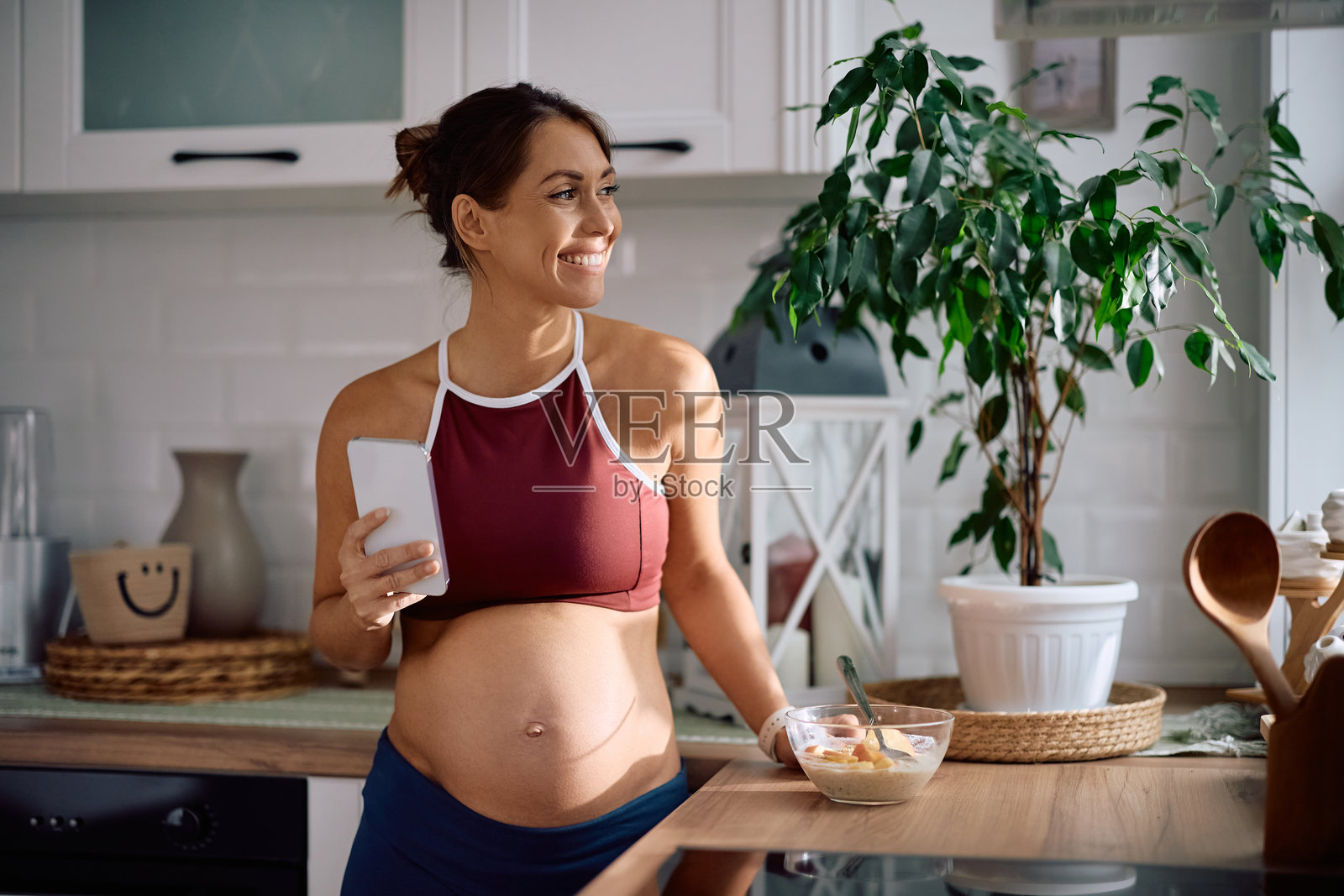 快乐的孕妇在厨房一边吃东西一边使用手机。照片摄影图片