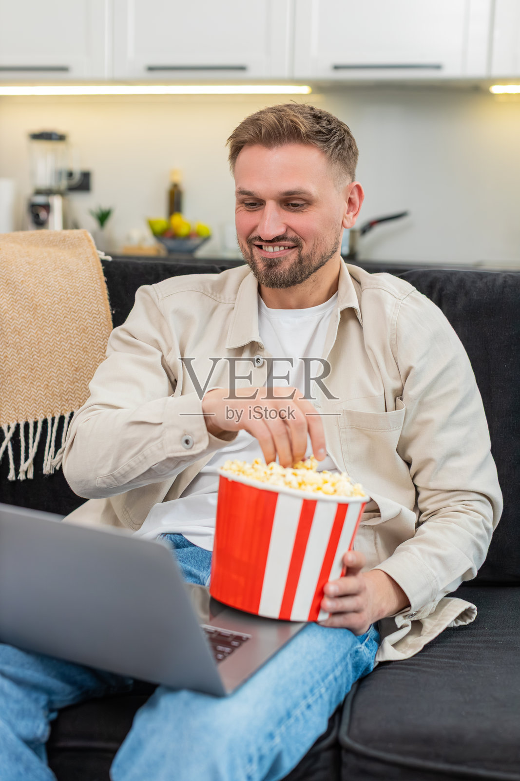 年轻男子在笔记本上看喜剧，手里拿着爆米花，开怀大笑，心情愉悦，享受有趣的夜晚时光照片摄影图片