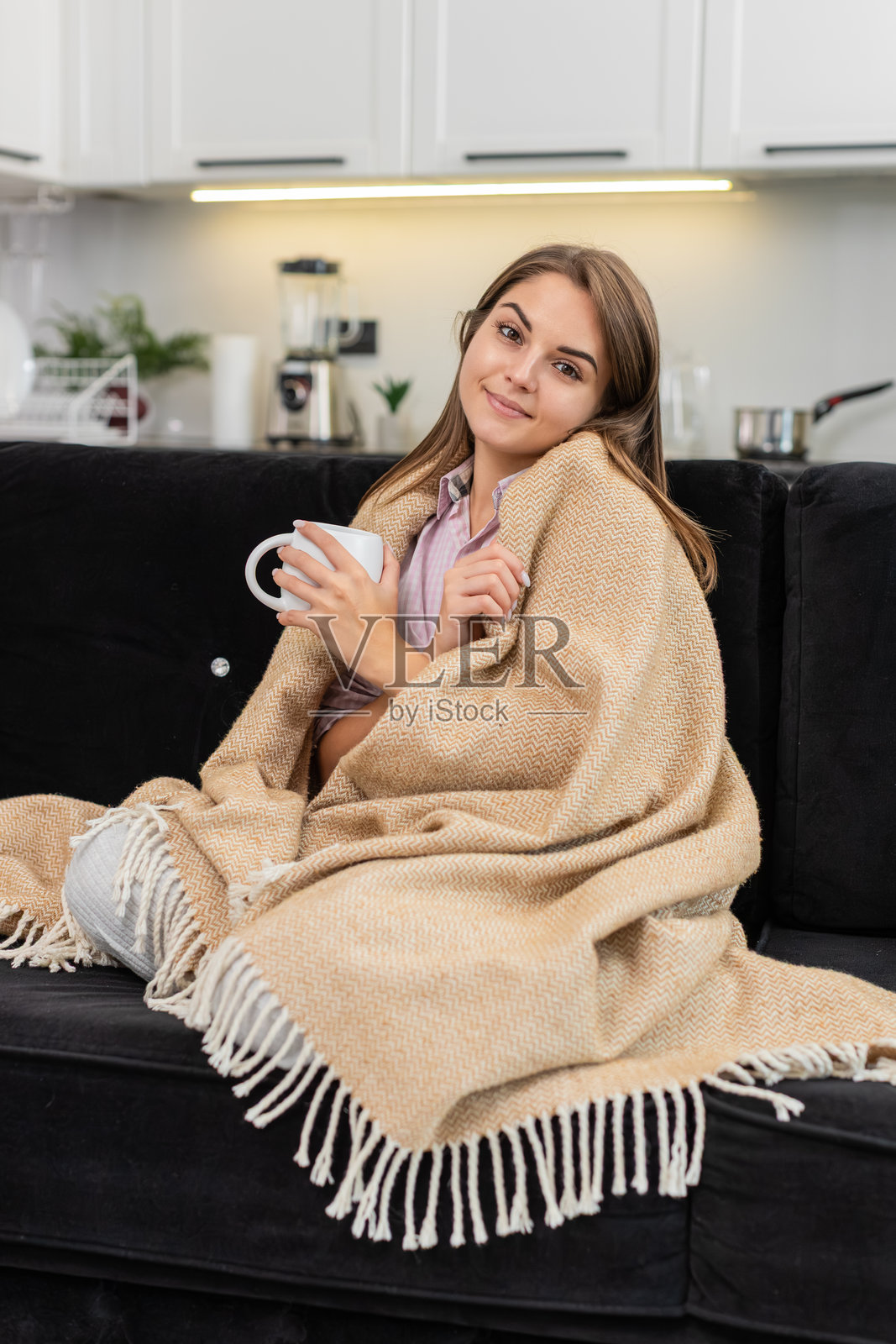 年轻女子抱着马克杯，在毯子下暖手，微笑着享受宁静的停顿和安详的深度休息照片摄影图片