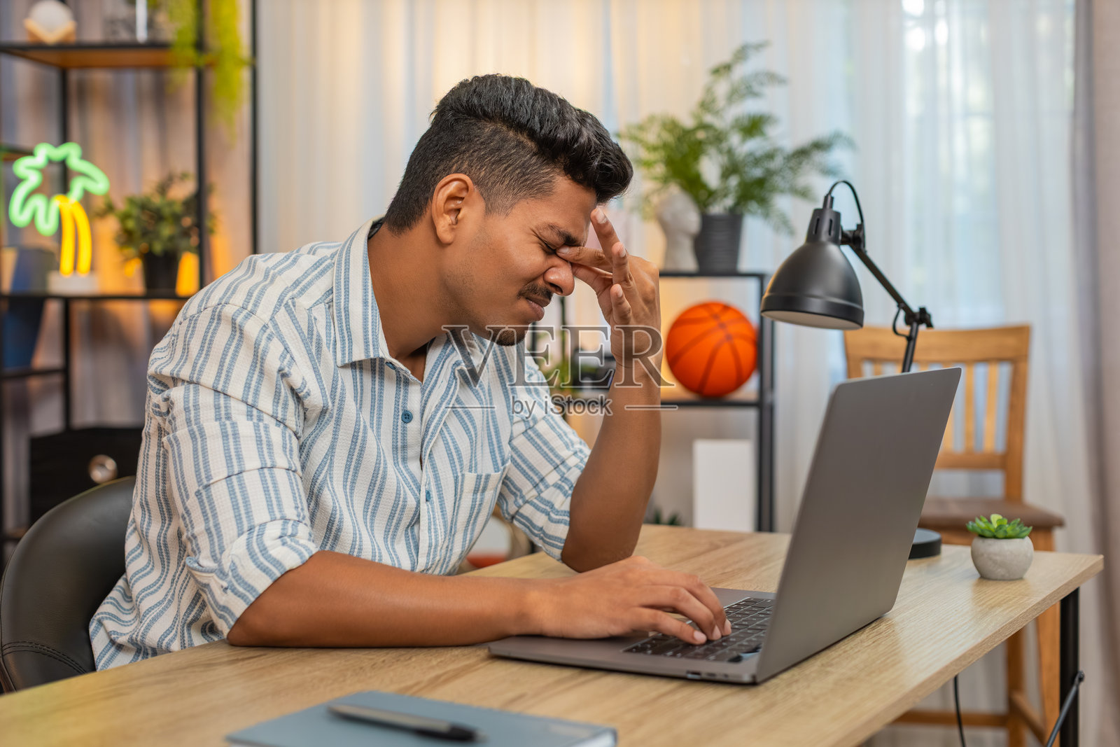 疲惫的印度男子在笔记本电脑上长时间工作后揉眼睛，感到眼部疲劳，需要在办公桌前休息片刻以缓解不适。照片摄影图片