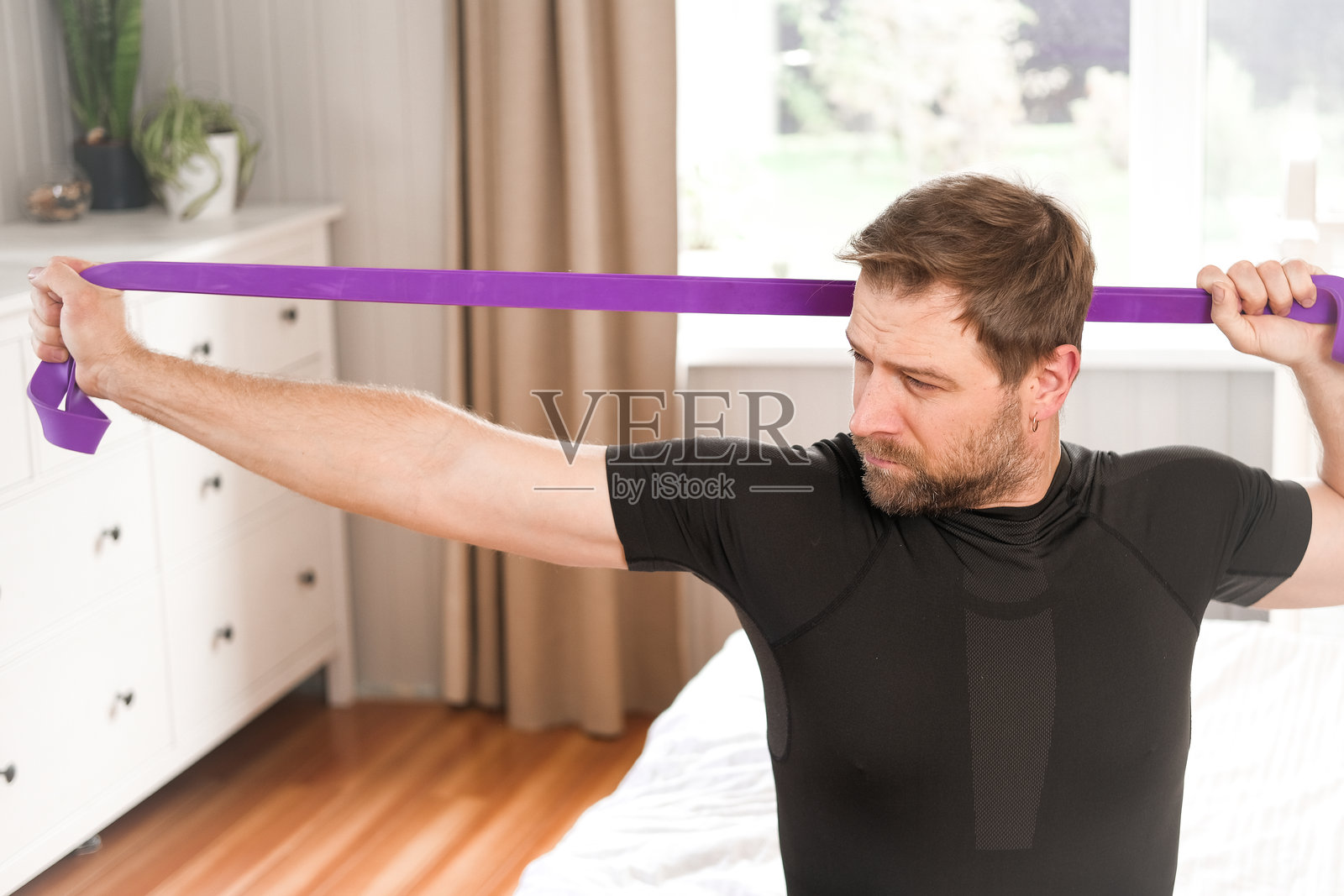 中年男性使用弹力带进行伸展。健康理念。40多岁的男性在家中的卧室进行颈部和肩部肌肉锻炼。拉伸以保持良好姿势。健康的生活方式，居家生活。照片摄影图片