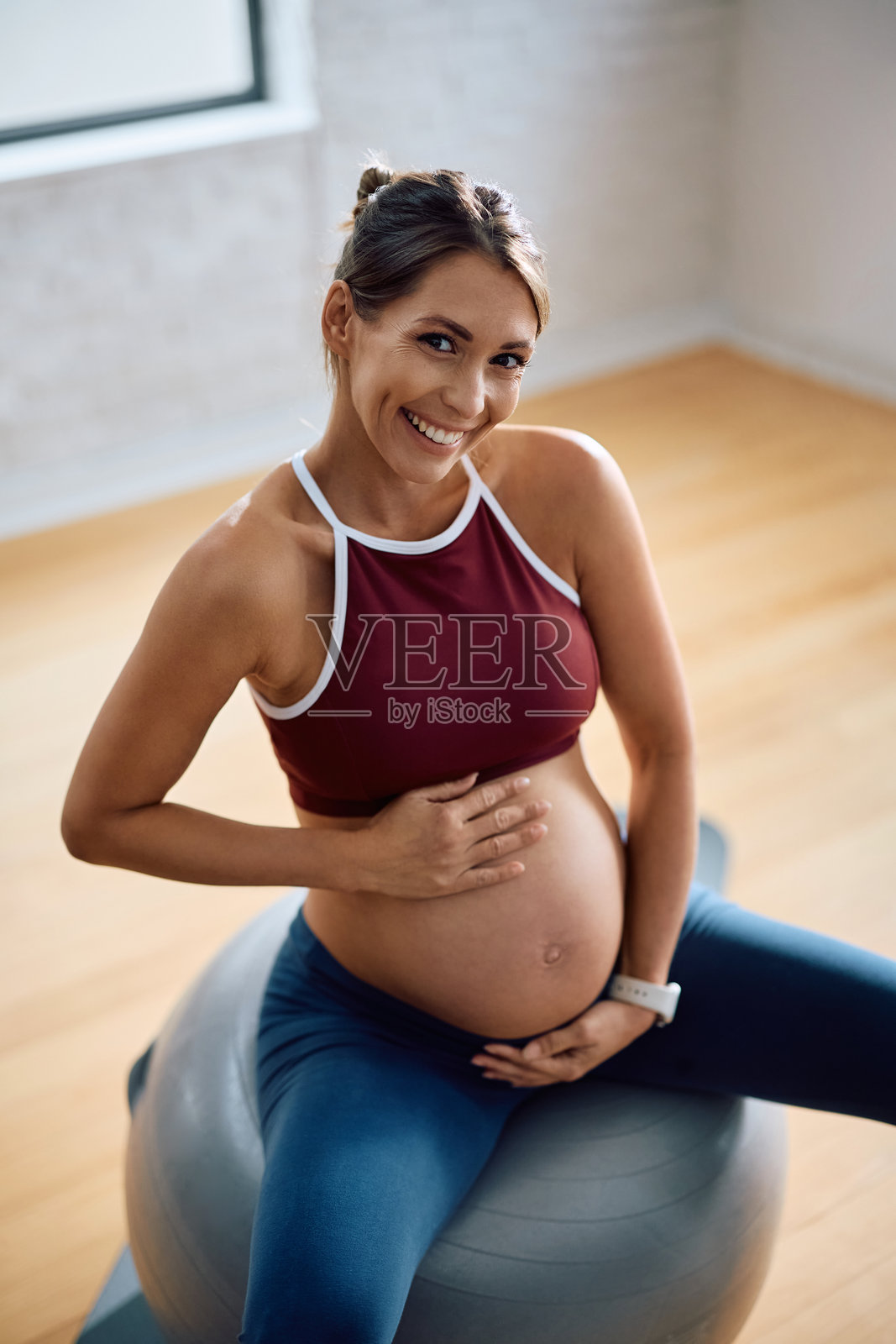 一位快乐的孕妇坐在瑜伽室的健身球上，一手扶着肚子，看向镜头。照片摄影图片