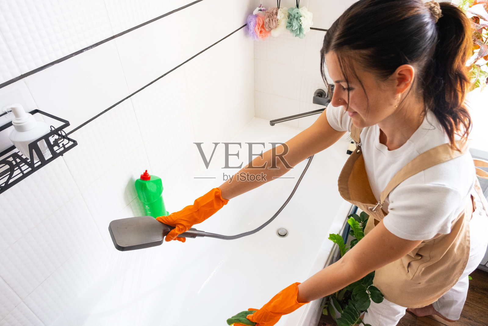 一位戴着橡胶手套、围着围裙的女士正在用海绵清洁剂和瓶装清洁剂手动清洗浴缸，浴缸位于房屋的浴室里。恢复家庭秩序、清洁和卫生，提供清洁服务。照片摄影图片