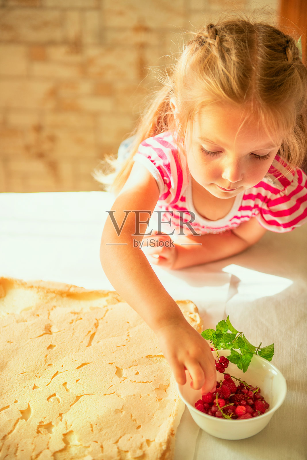 小女孩在装饰蛋白卷的同时，伸手去拿碗里新鲜的覆盆子。体现了童年、烹饪、健康饮食和家庭传统等概念。照片摄影图片