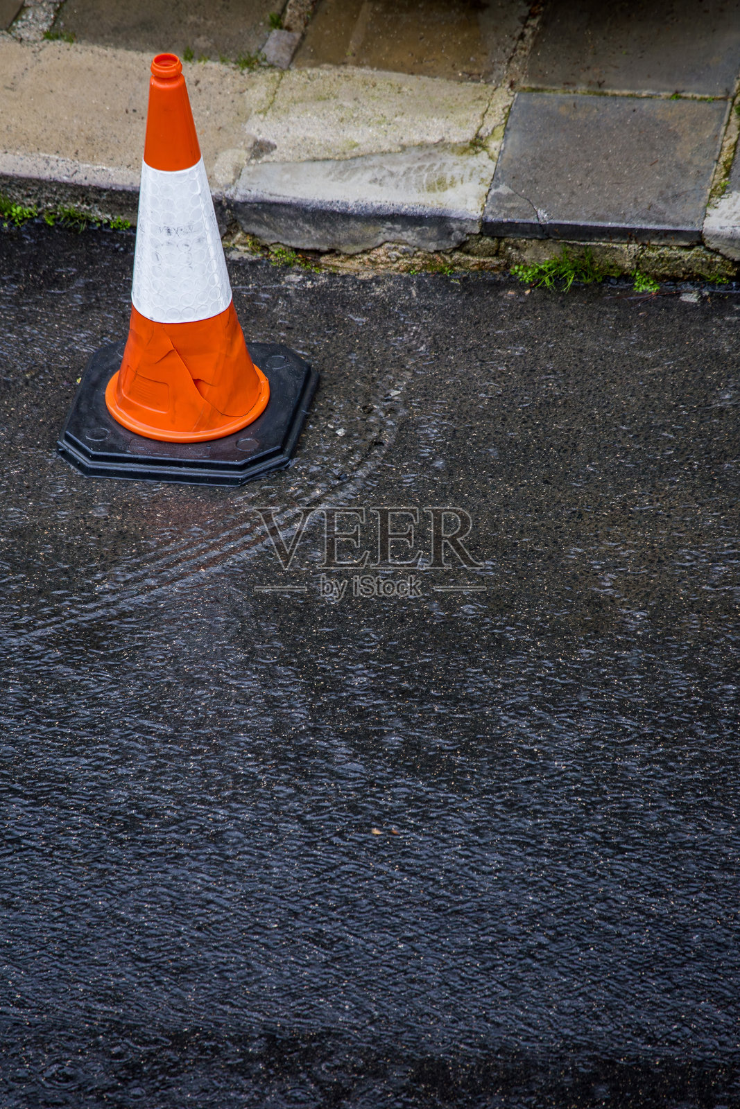 一个橙白相间的交通锥立在潮湿的街道旁，靠近路缘，雨水在其底部激起涟漪。这个高角度的视角捕捉到了雨天城市宁静而略带忧郁的氛围。照片摄影图片