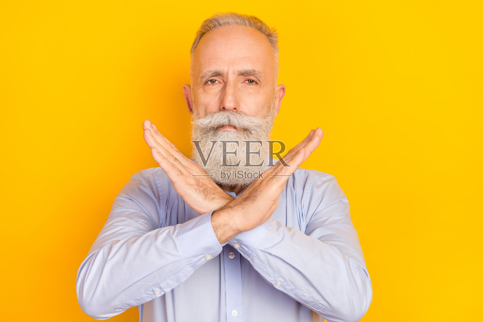 照片：蓄着胡须的严肃中年男子身穿蓝色衬衫，双臂交叉，背景为黄色照片摄影图片