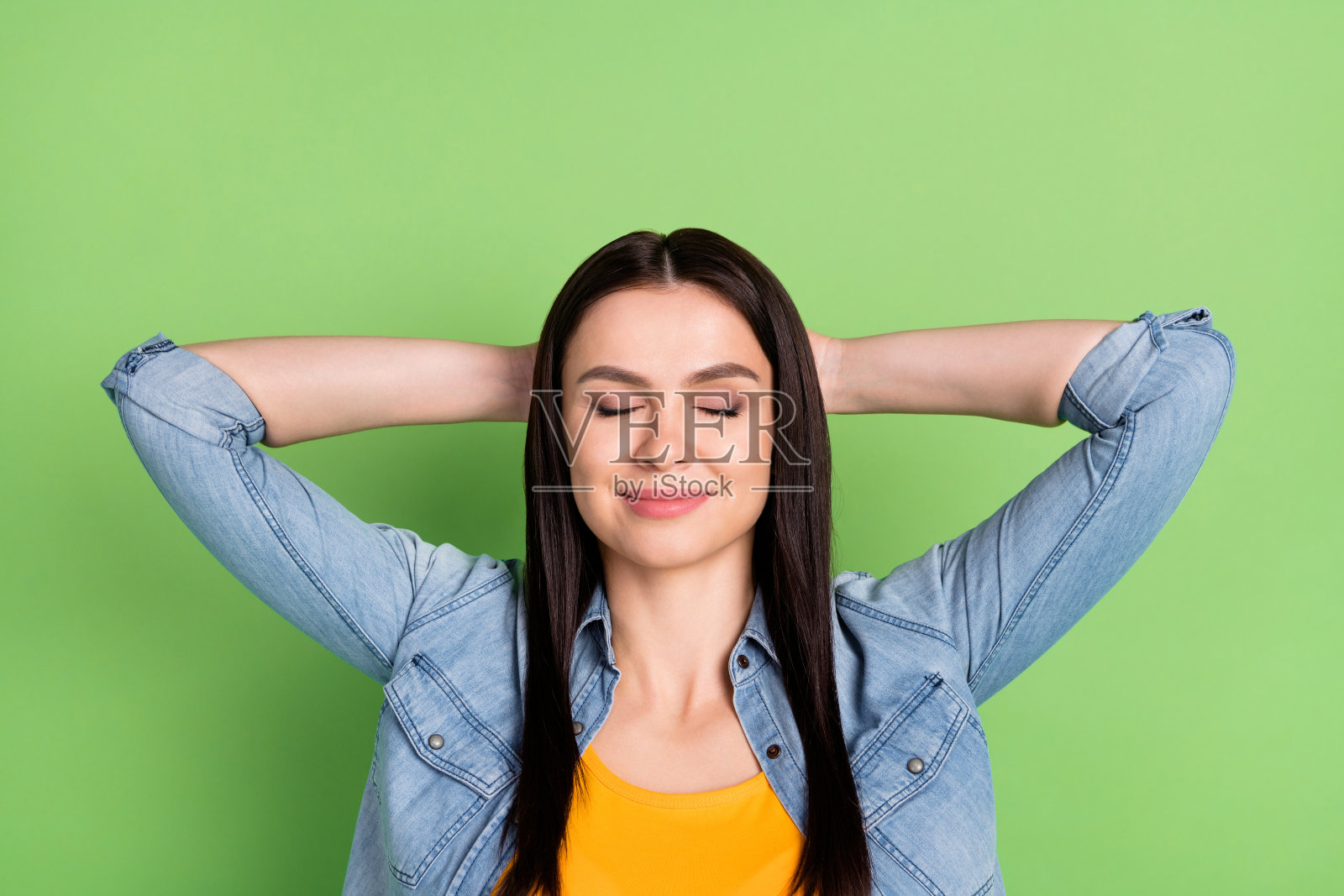 照片：一位乐观的棕色长发女士，双手搭在头上，身穿牛仔衬衫，孤立在粉绿色背景上照片摄影图片
