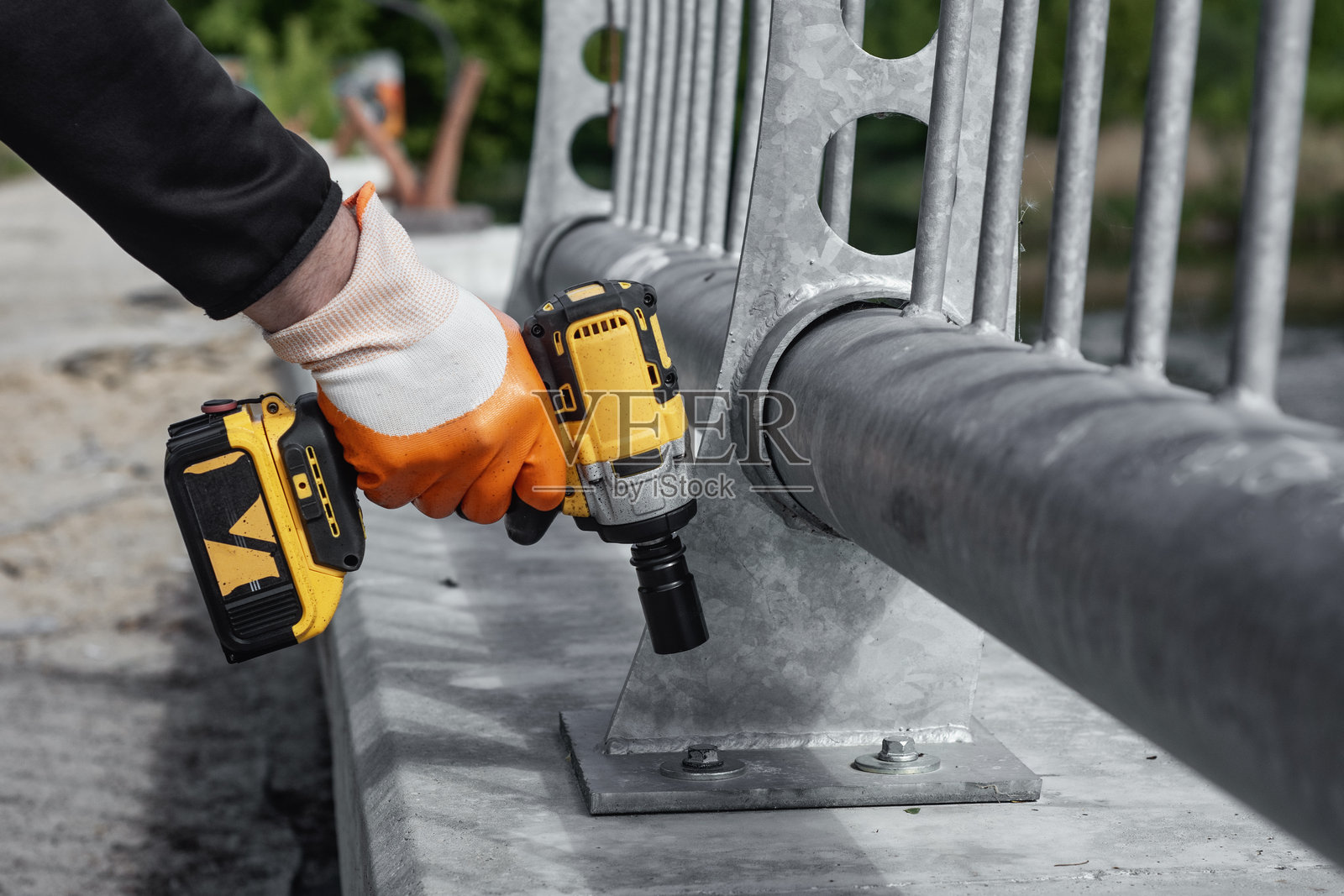 一名身穿安全背心和橙色手套的工人在建筑工地上手持电钻，靠近金属栏杆。概念是建筑工具、安全装备和工作现场设备。照片摄影图片