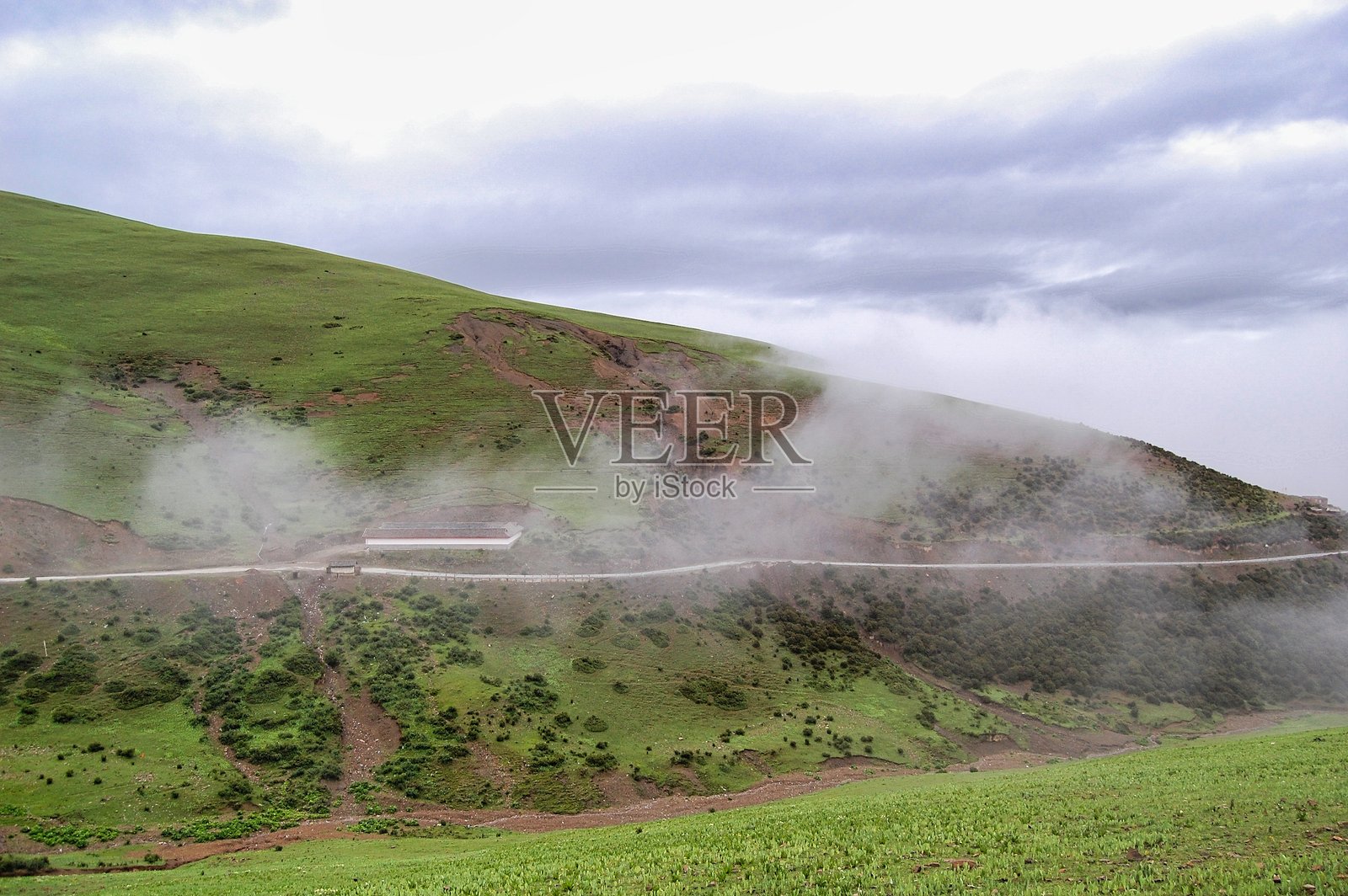云雾缭绕的山路，两侧是绿色的山坡，天空布满云彩。照片摄影图片