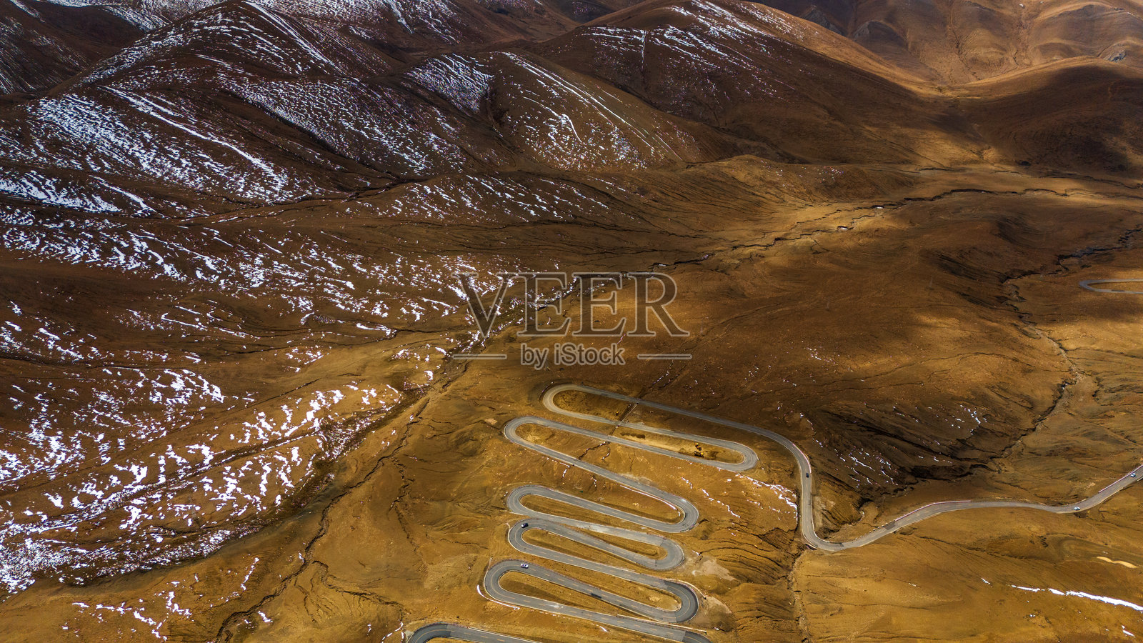 从藏区一侧俯瞰通往珠穆朗玛峰的蜿蜒道路，周围是崎岖的棕色山脉和零星的雪地，天空戏剧性地变幻着。照片摄影图片