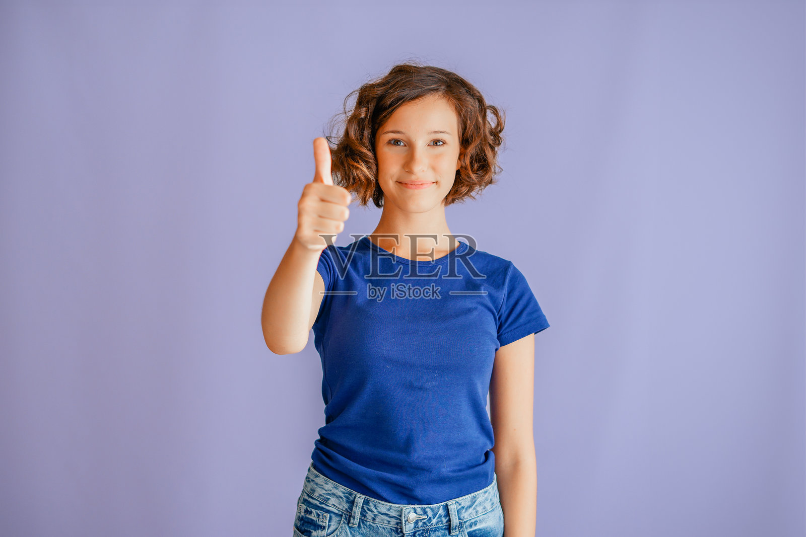 一位迷人的年轻女子带着自信的微笑和积极的能量竖起大拇指，在充满活力的蓝色工作室背景下，营造出一种愉悦的氛围，传递着赞同的理念。照片摄影图片