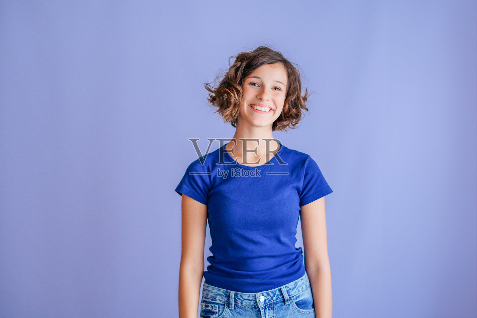 穿着蓝色T恤、留着波波头卷发的年轻女子，脸上带着快乐友好的笑容，对着镜头，背景是充满活力的蓝色影棚。照片摄影图片