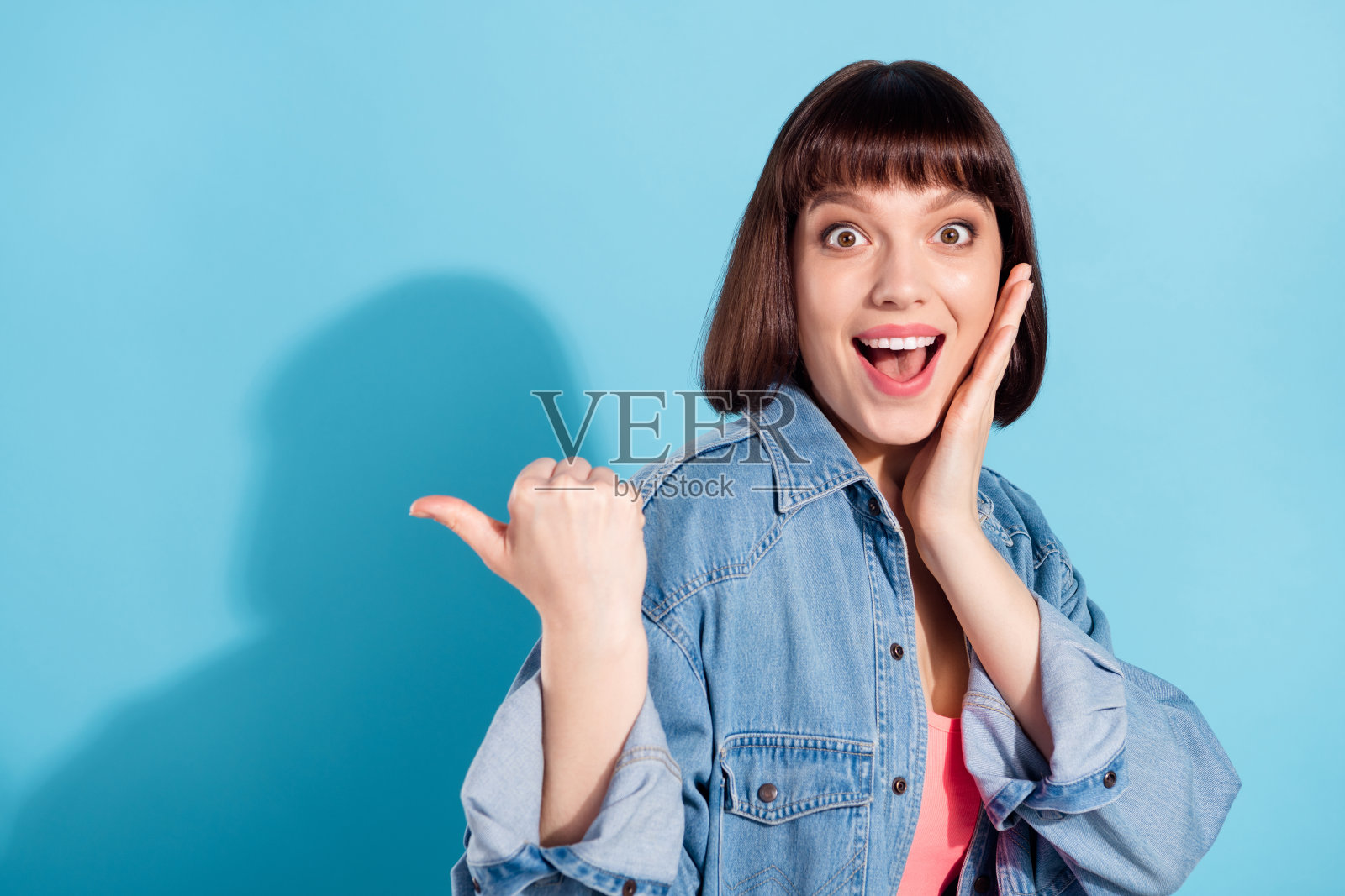 梦幻般印象深刻的年轻女子，身穿牛仔衬衫，微笑，手搭脸颊，手指指向后方空白处，孤立在蓝色背景上。照片摄影图片