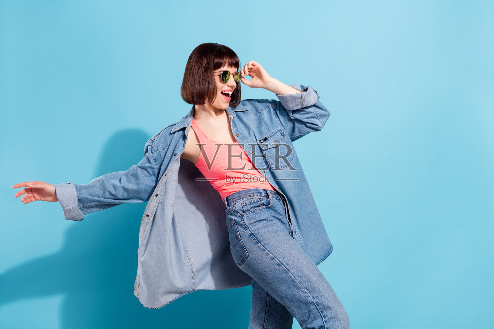 照片：时尚迷人的年轻女子，身穿牛仔衬衫，戴着深色眼镜，面带笑容，在蓝色的背景前跳舞。照片摄影图片