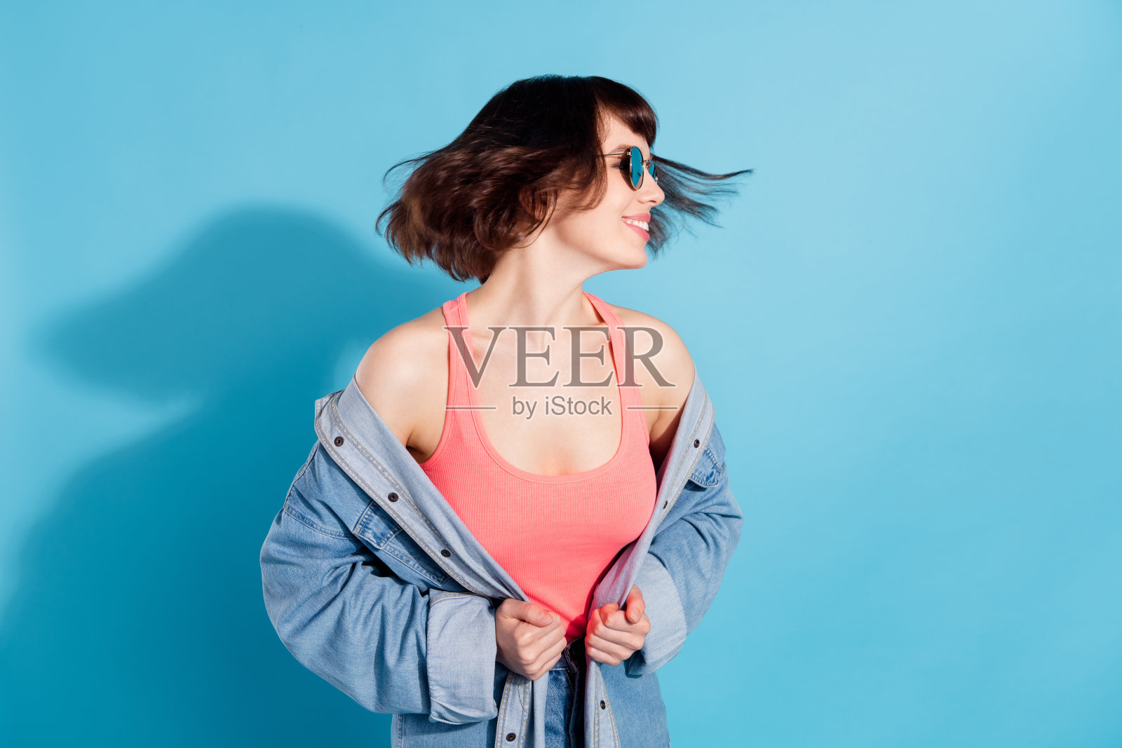 身穿牛仔衬衫、戴着墨镜的年轻女士，笑容灿烂，在蓝色背景前跳舞，充满活力，可爱时尚照片摄影图片