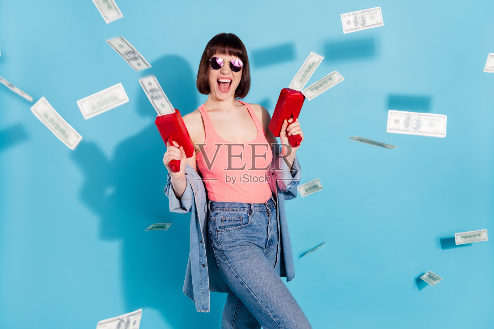 身穿粉色上衣、牛仔裤和眼镜的疯狂年轻女子，手持钱枪，在蓝色背景前大喊大叫，孤立拍摄照片摄影图片