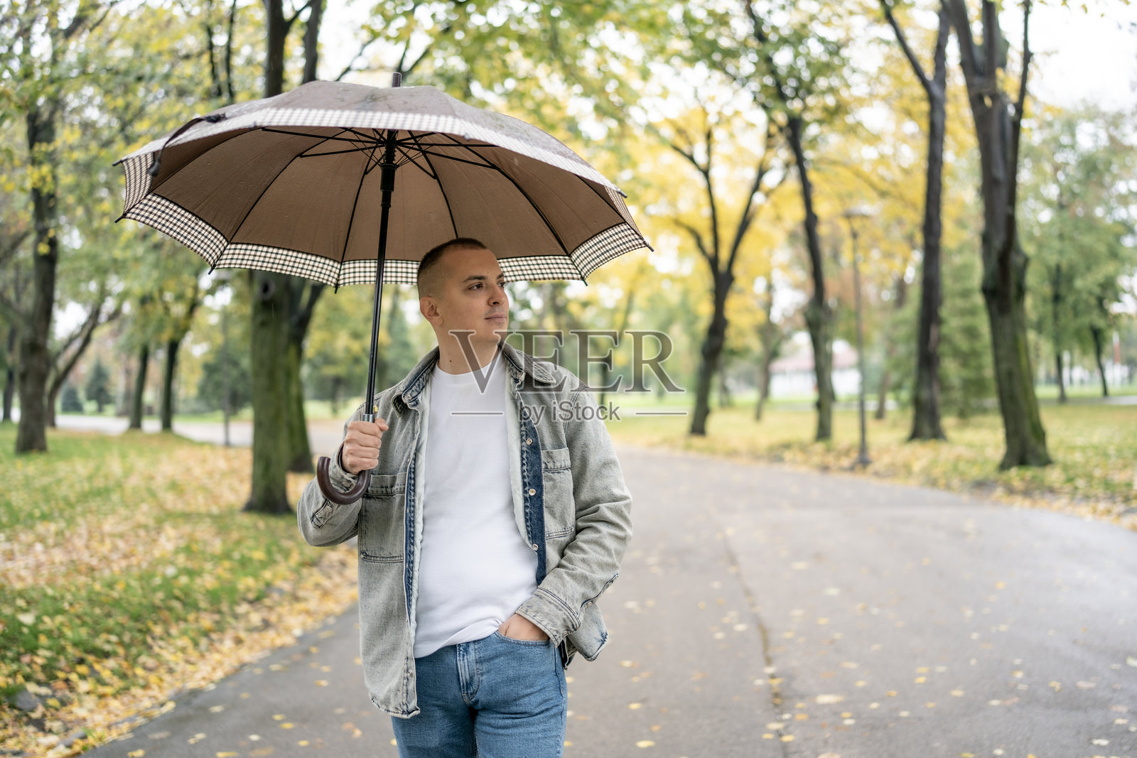一个男人穿着休闲牛仔裤和牛仔夹克，雨天打着伞走在秋天的公园里照片摄影图片
