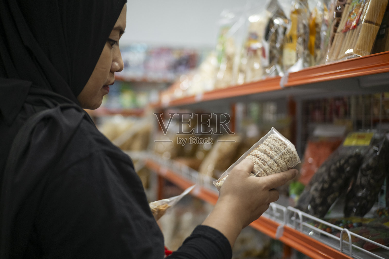 一位穿着头巾和端庄服装的年轻美丽的印度尼西亚女性，周末在超市货架上购物并购买零食。照片摄影图片