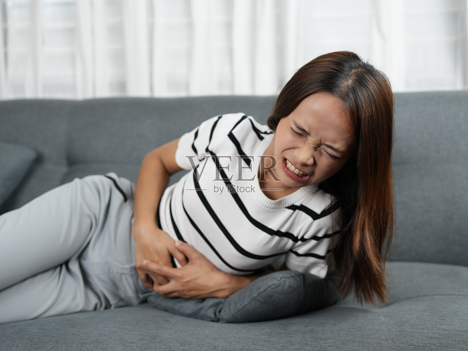 女子坐在沙发上，面露痛苦之色，腹部疼痛难忍，背景是灯光柔和的舒适客厅，突显她的不适感。照片摄影图片