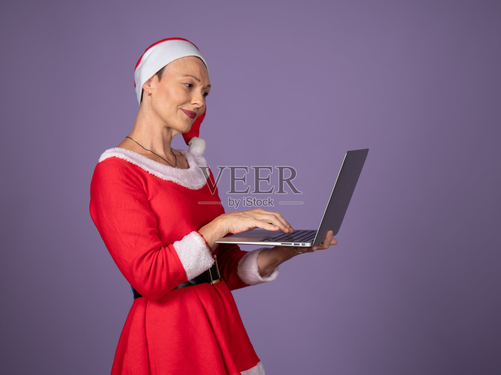 身穿圣诞老人服装的女士正在使用笔记本电脑，背景是圣诞节的场景。照片摄影图片