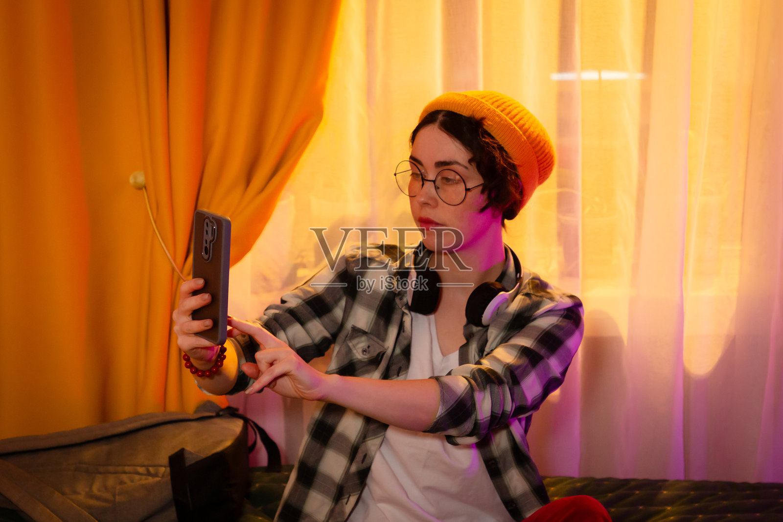 穿着休闲服、戴着绒线帽和耳机的女孩躺在床上放松并自拍。背景是日落余晖和舒适的居家环境。中景拍摄。照片摄影图片