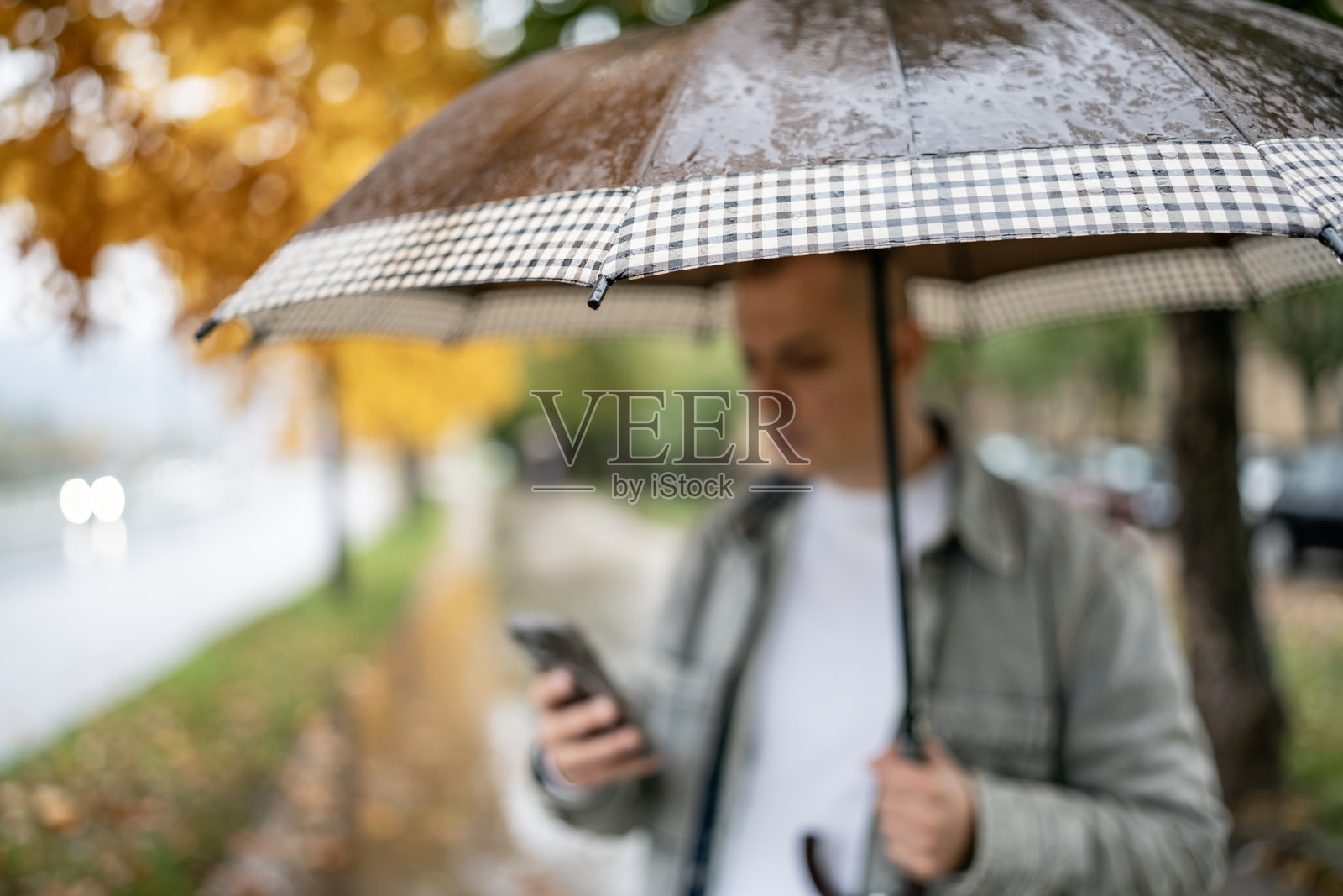 手持电话的男子在雨天的秋街上撑着格子伞行走照片摄影图片