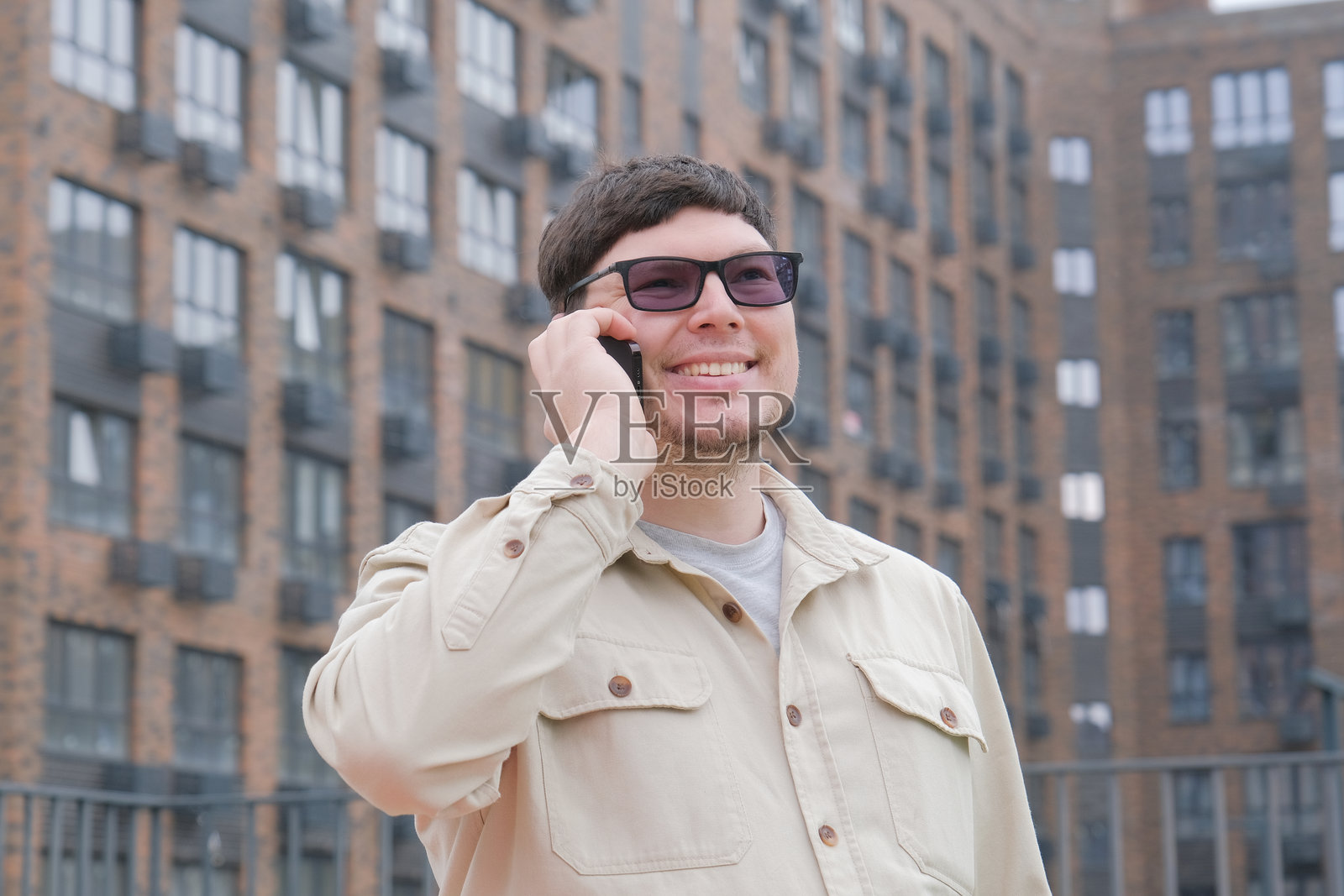 一个穿着休闲服的男人，站在户外，身前是大型公寓楼，他正在打电话。照片摄影图片