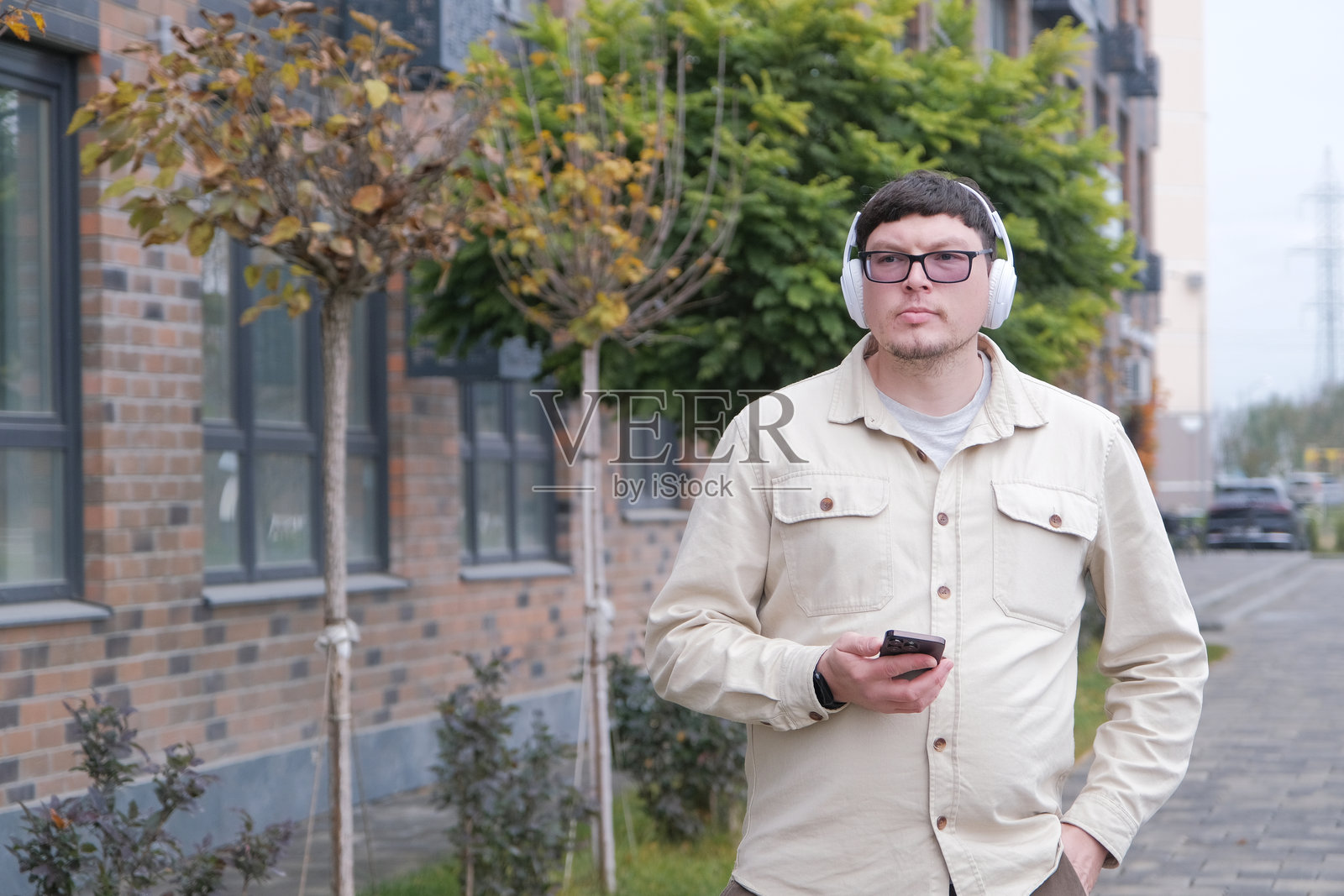 一个人戴着耳机，使用智能手机听音乐，走在公寓楼旁照片摄影图片