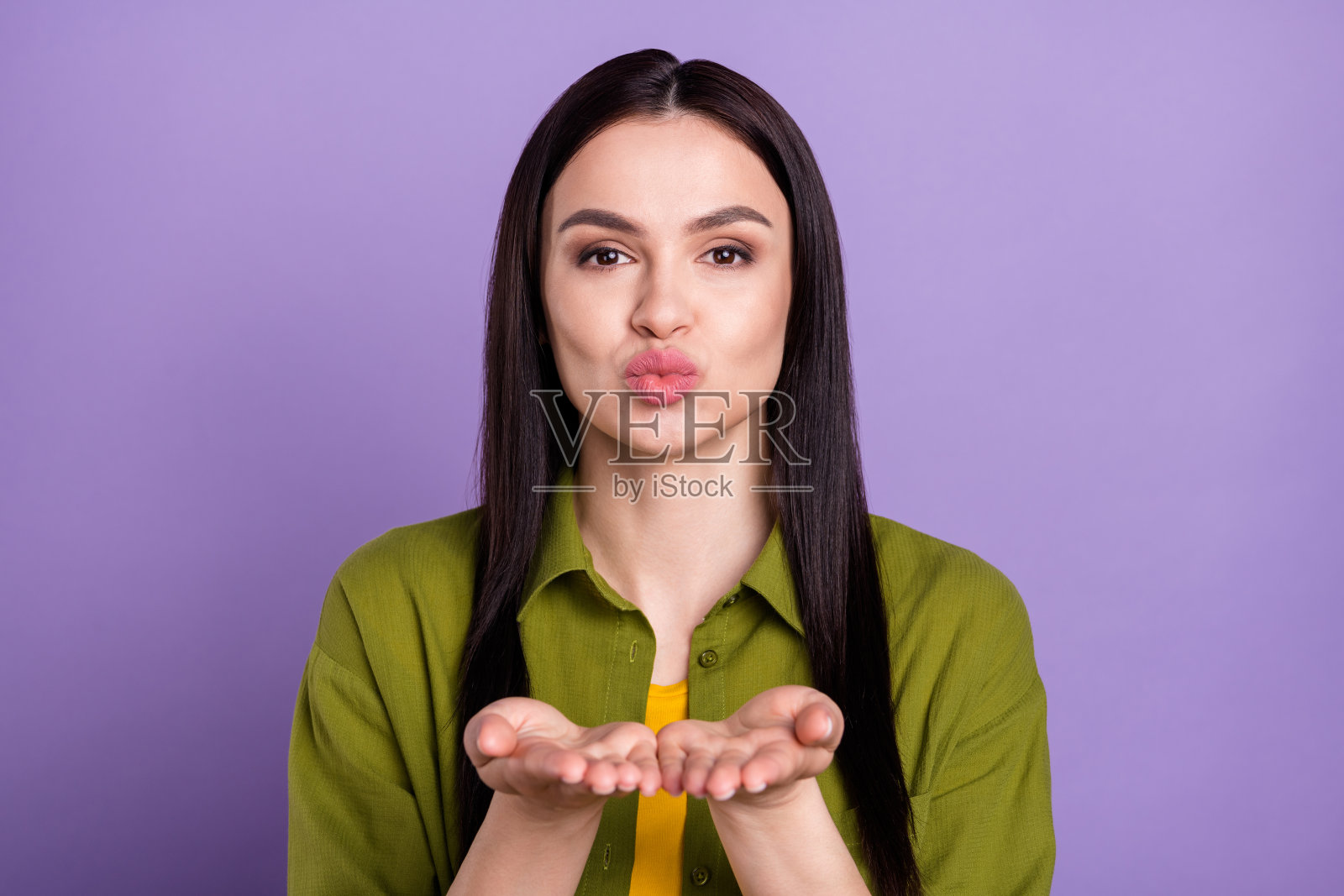 身穿卡其色服装的漂亮闪亮年轻女子，嘴唇噘起，亲吻你的手臂，孤立在紫色背景上照片摄影图片