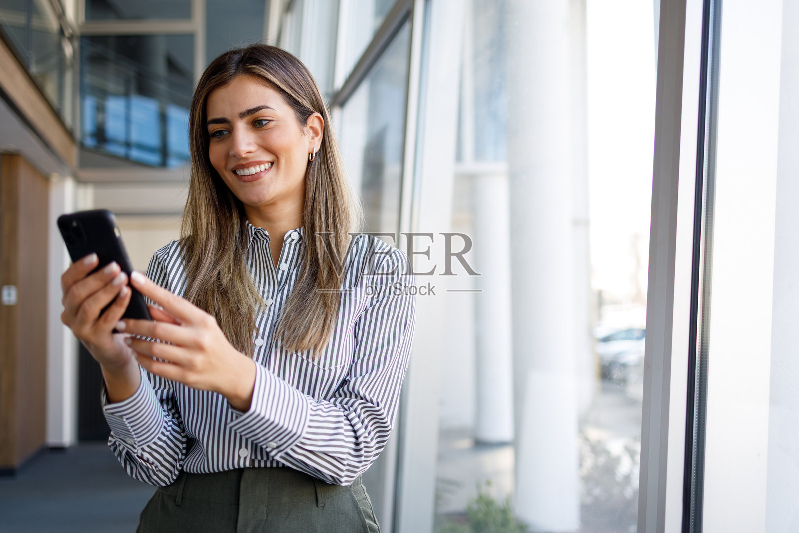 一位职业女性在现代办公楼内微笑着，用手机交流，享受成功的职业生涯照片摄影图片