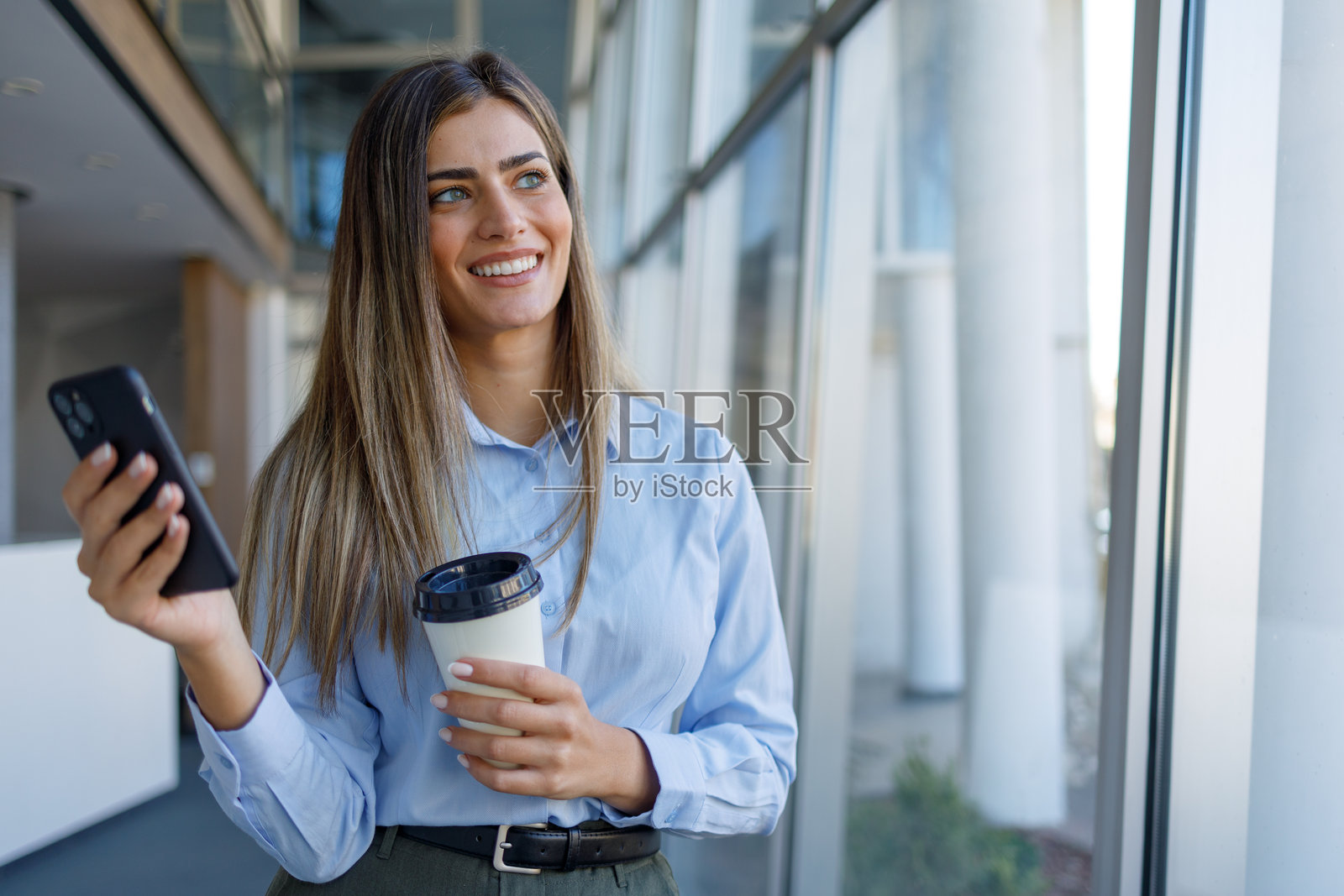 一位女商人在现代办公室走廊里，一手拿着智能手机和咖啡杯，目光望向别处。照片摄影图片