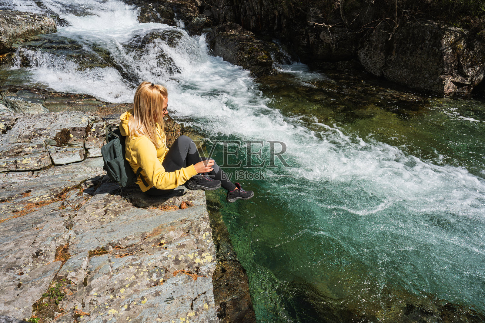 一位背着背包的女游客在挪威湍急的山间河流旁的一块岩石上休息。照片摄影图片