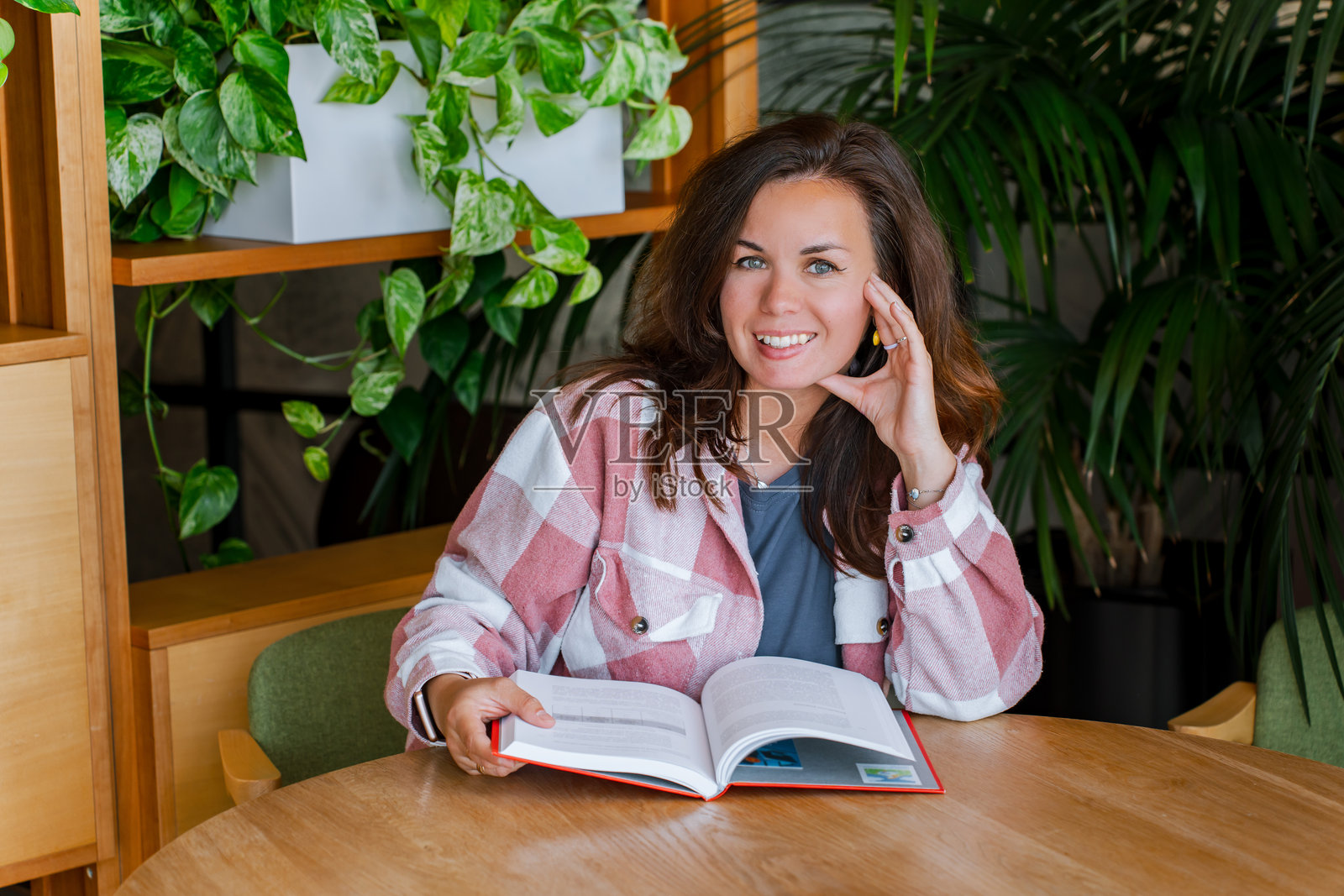 一位年轻女子穿着衬衫，手捧书籍正在学习，一个学生坐在一张木桌旁看书，室内有绿植。照片摄影图片