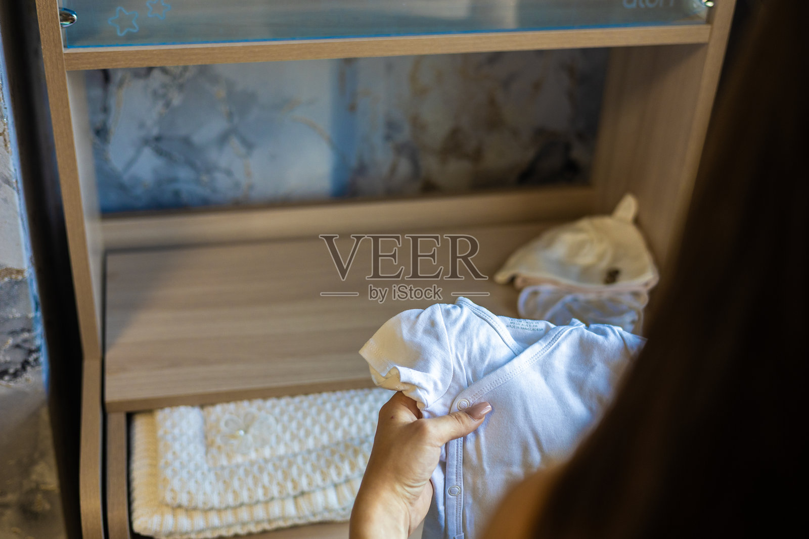 一位孕妇正小心翼翼地拿着一件白色婴儿连体衣，在现代化的婴儿房里整理衣物和配饰，为即将到来的新生儿做准备。照片摄影图片