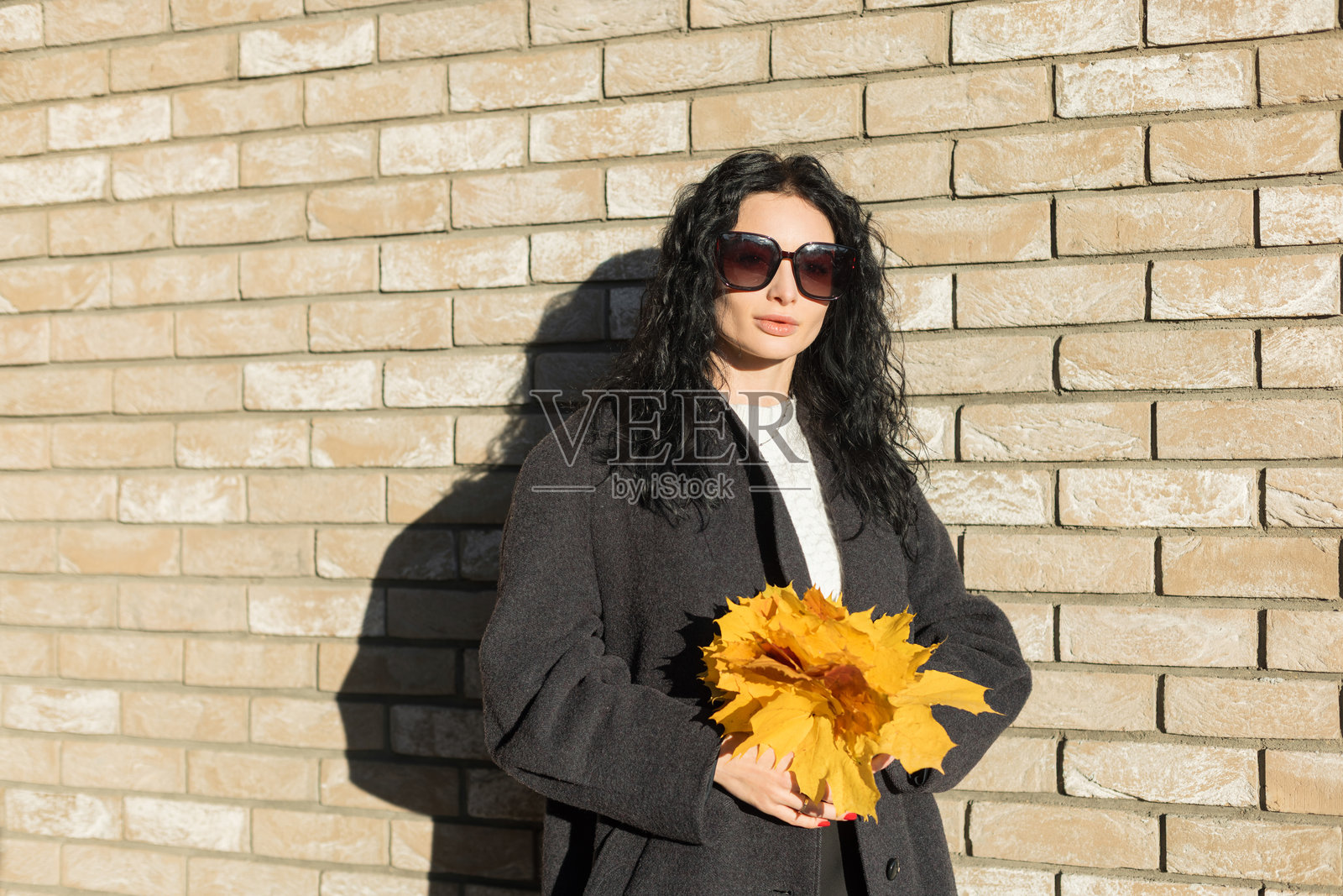 一位卷发女子在质朴的砖墙背景前，面带温暖的微笑，手持明亮的黄色枫叶，拍摄的秋季肖像。照片摄影图片