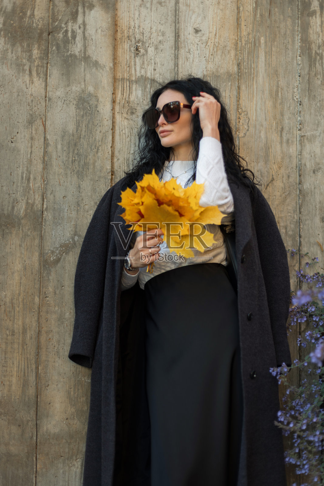 留着卷曲黑发的漂亮女子，身着秋季服装，手持黄叶，倚靠在木墙边。季节性肖像与秋季时尚灵感。照片摄影图片