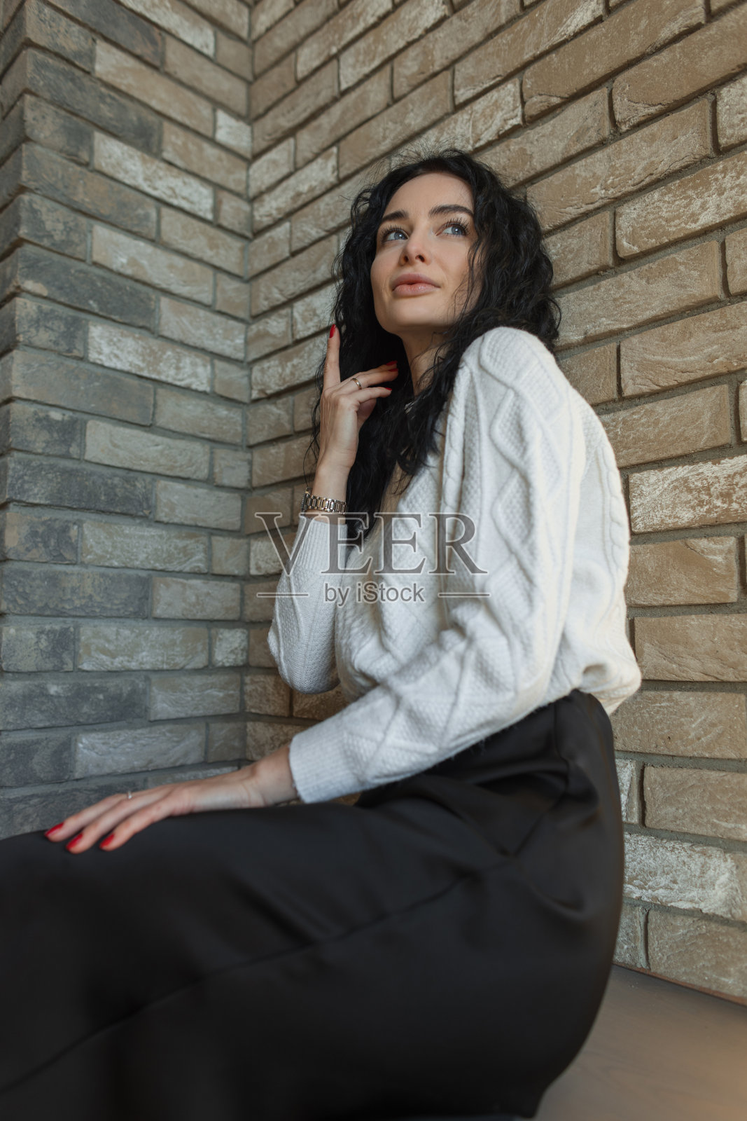 一位现代女性穿着时尚的秋季服装，在城市砖墙旁户外休息，显得放松而自信。这位女孩穿着舒适的针织毛衣和裙子，坐在温暖的阳光下。照片摄影图片