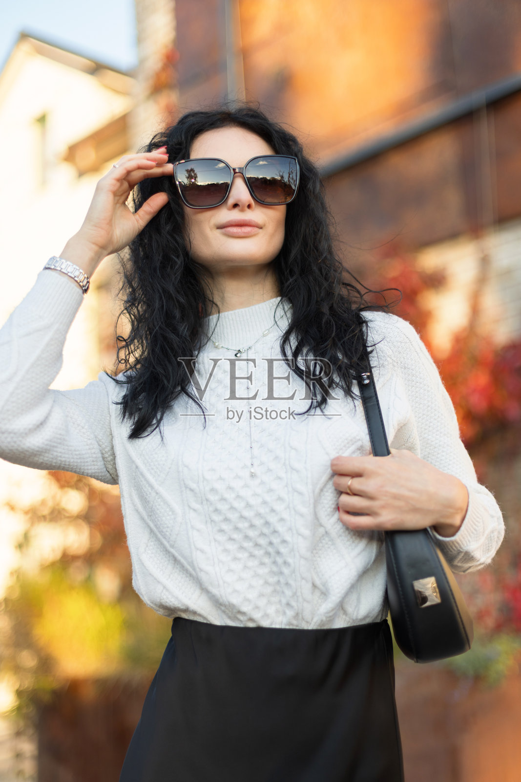 戴着太阳镜的优雅女士在红叶环绕的砖墙旁摆姿势。她身着时尚的秋季服装，包括白色毛衣和黑色裙子，在户外享受温暖的季节阳光。照片摄影图片