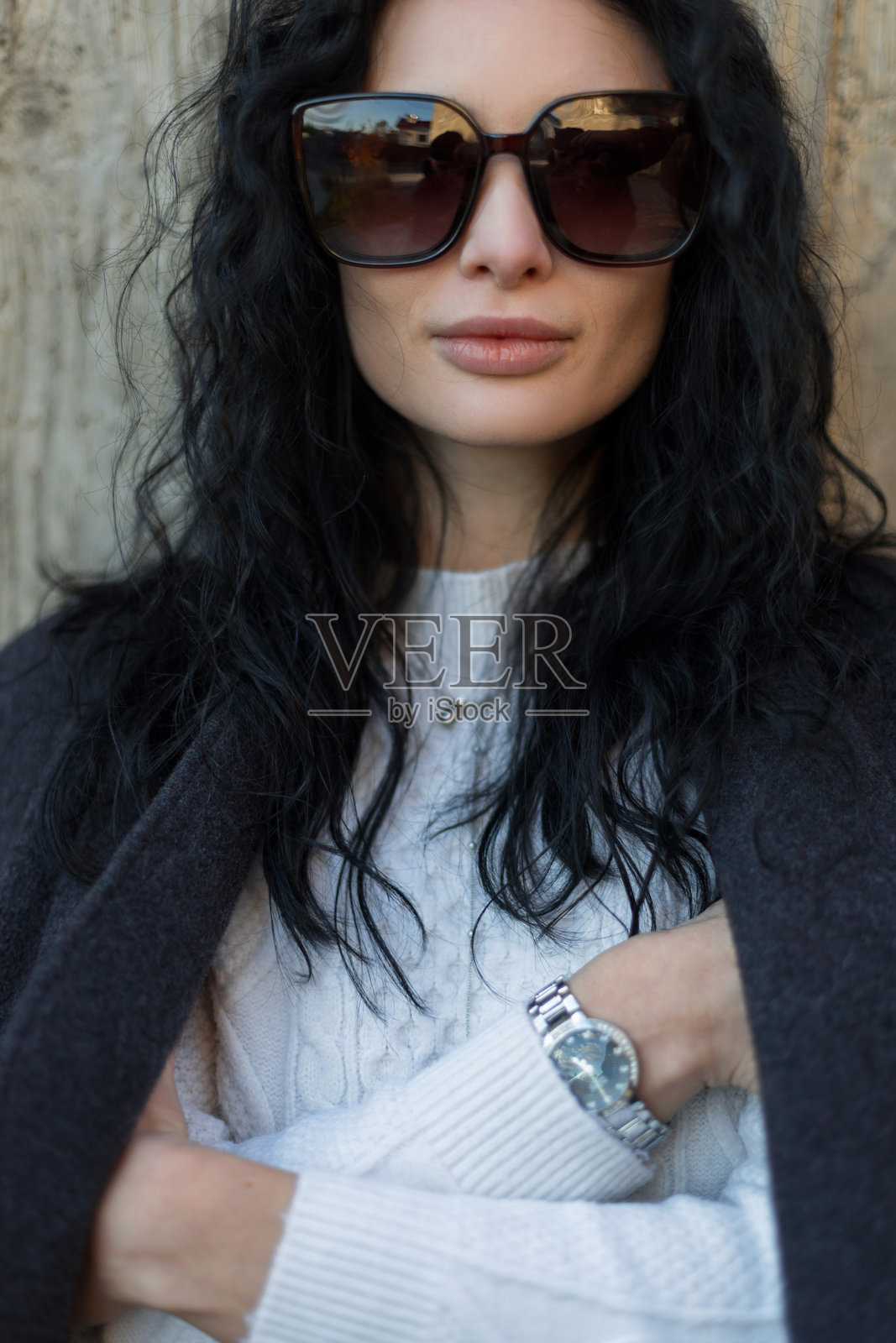 一位优雅的女士，留着黑色卷发，戴着太阳镜，身穿灰色外套，在户外的水泥墙旁摆姿势。秋季时尚，休闲街头风格，现代女性肖像概念。照片摄影图片