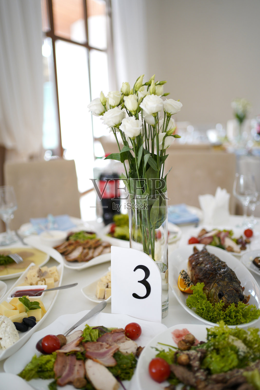 节日餐桌上摆放着各种菜肴、沙拉、肉类和鱼类，餐桌上还装饰着花瓶里的鲜花和桌号。照片摄影图片