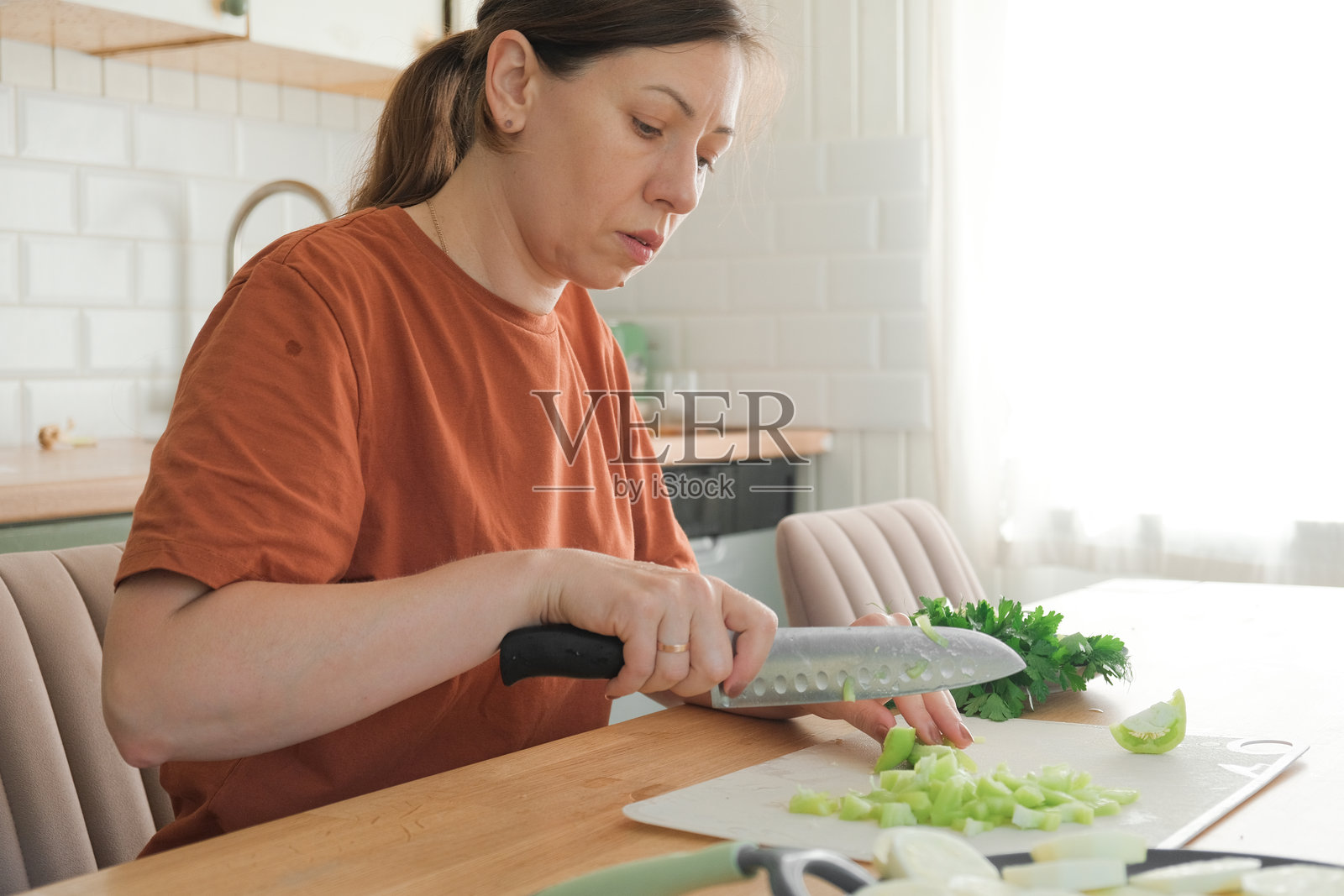 中年女性穿着陶土色T恤，在自家厨房准备绿色新鲜的纯素沙拉。她正拿着刀在砧板上切蔬菜和生菜，烹饪餐食。健康的饮食食物。照片摄影图片