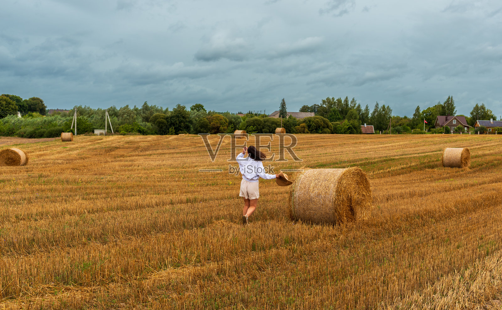 女子走过收割后的田野，田野里堆放着干草垛照片摄影图片